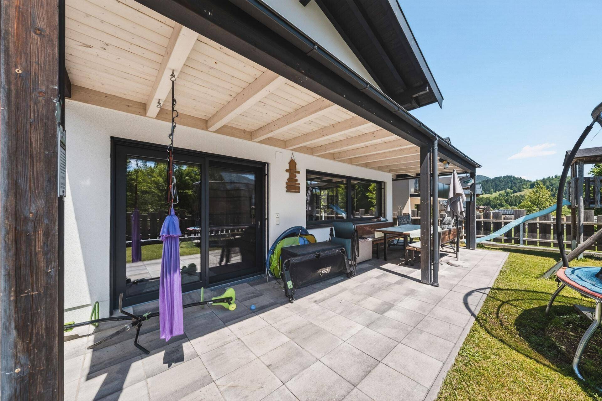 Überdachte Terrasse mit Sitzgelegenheiten und Zugang zum Garten mit Spielgeräten.