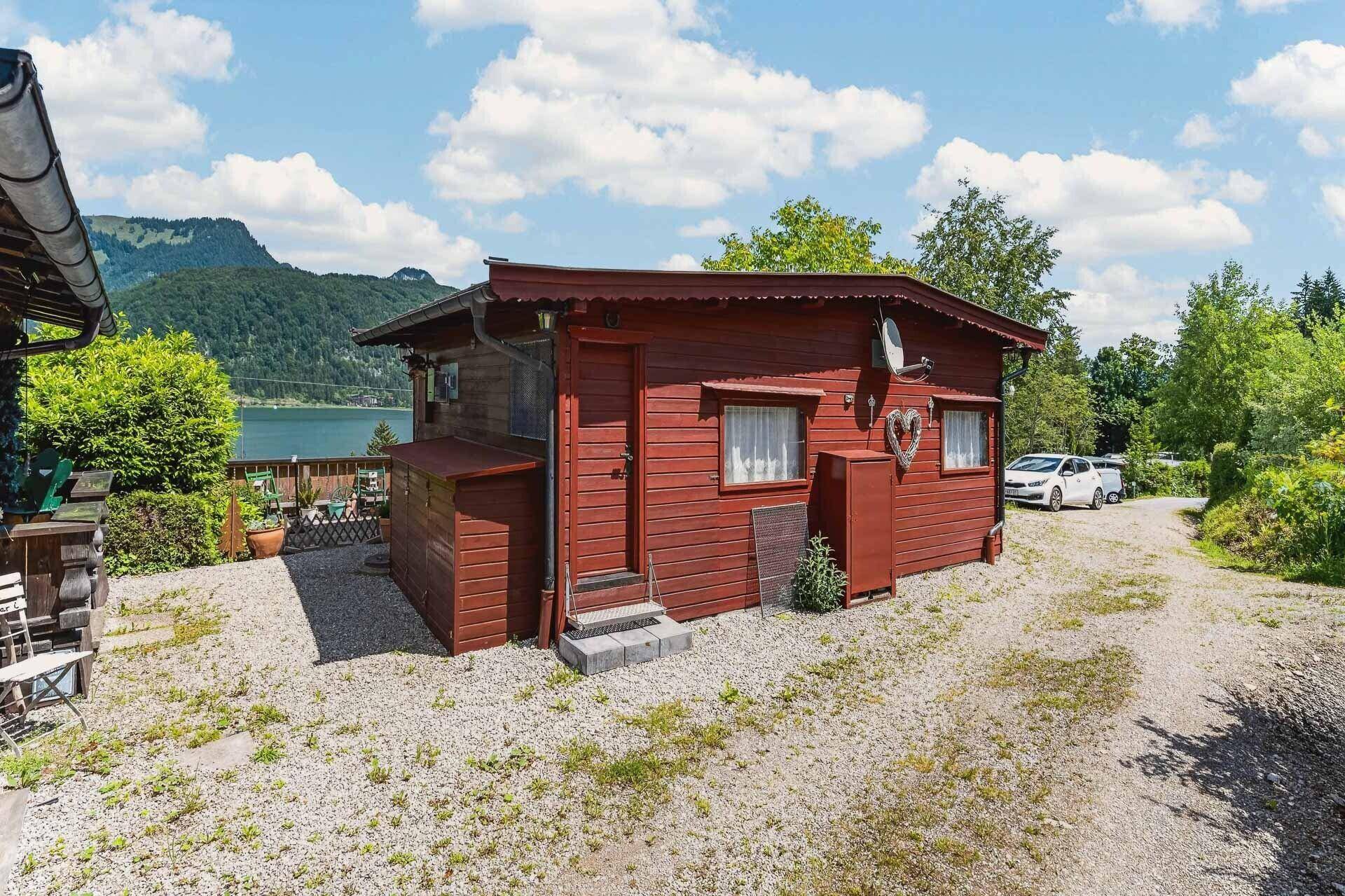 Rotes Holzhaus mit Schotterfläche und Blick auf den See im Hintergrund, umgeben von Bäumen.