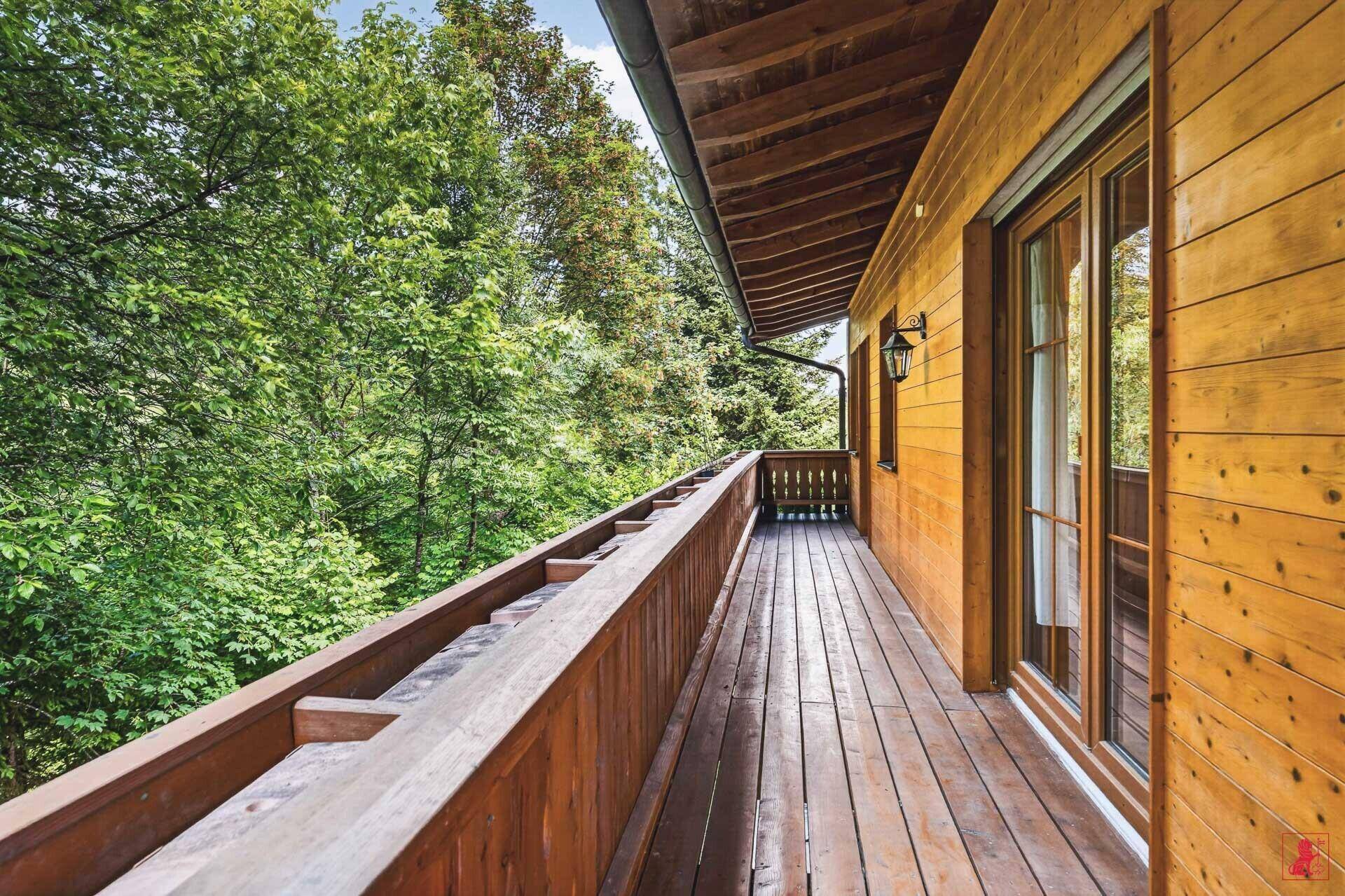 Breiter Holzbalkon entlang der Hausseite, umgeben von üppigem Grün und Bäumen.