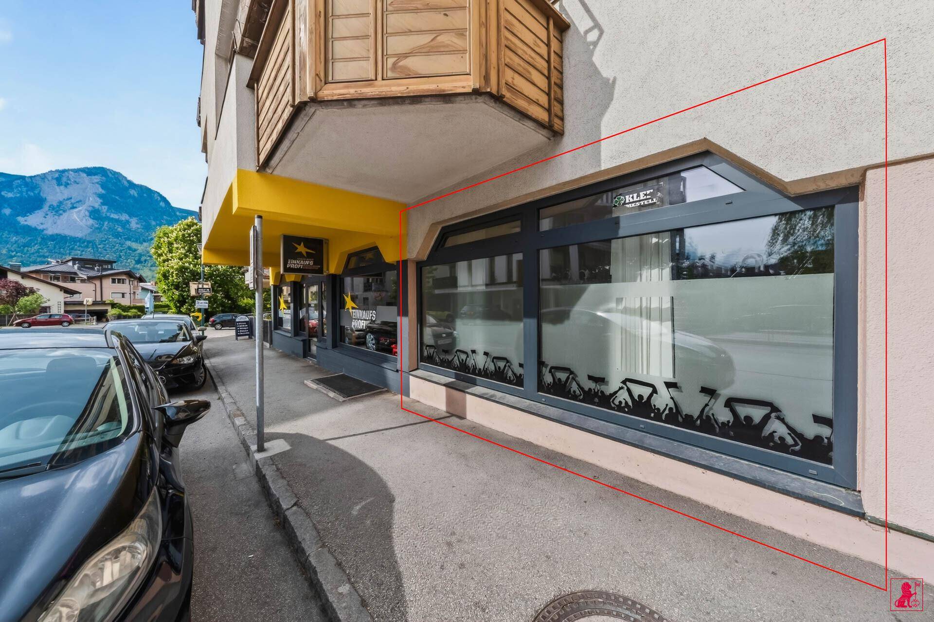 Außenansicht der Geschäftsfassade mit großen Fenstern, geparkten Autos und Bergen im Hintergrund.
