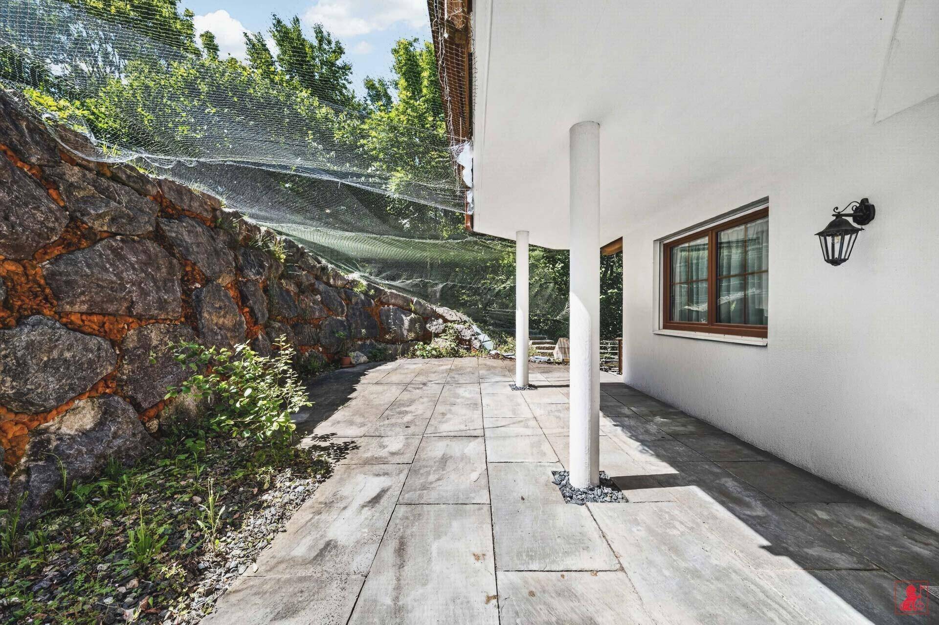 Überdachte Terrasse mit Steinplatten und Säulen, umgeben von einer Natursteinmauer.