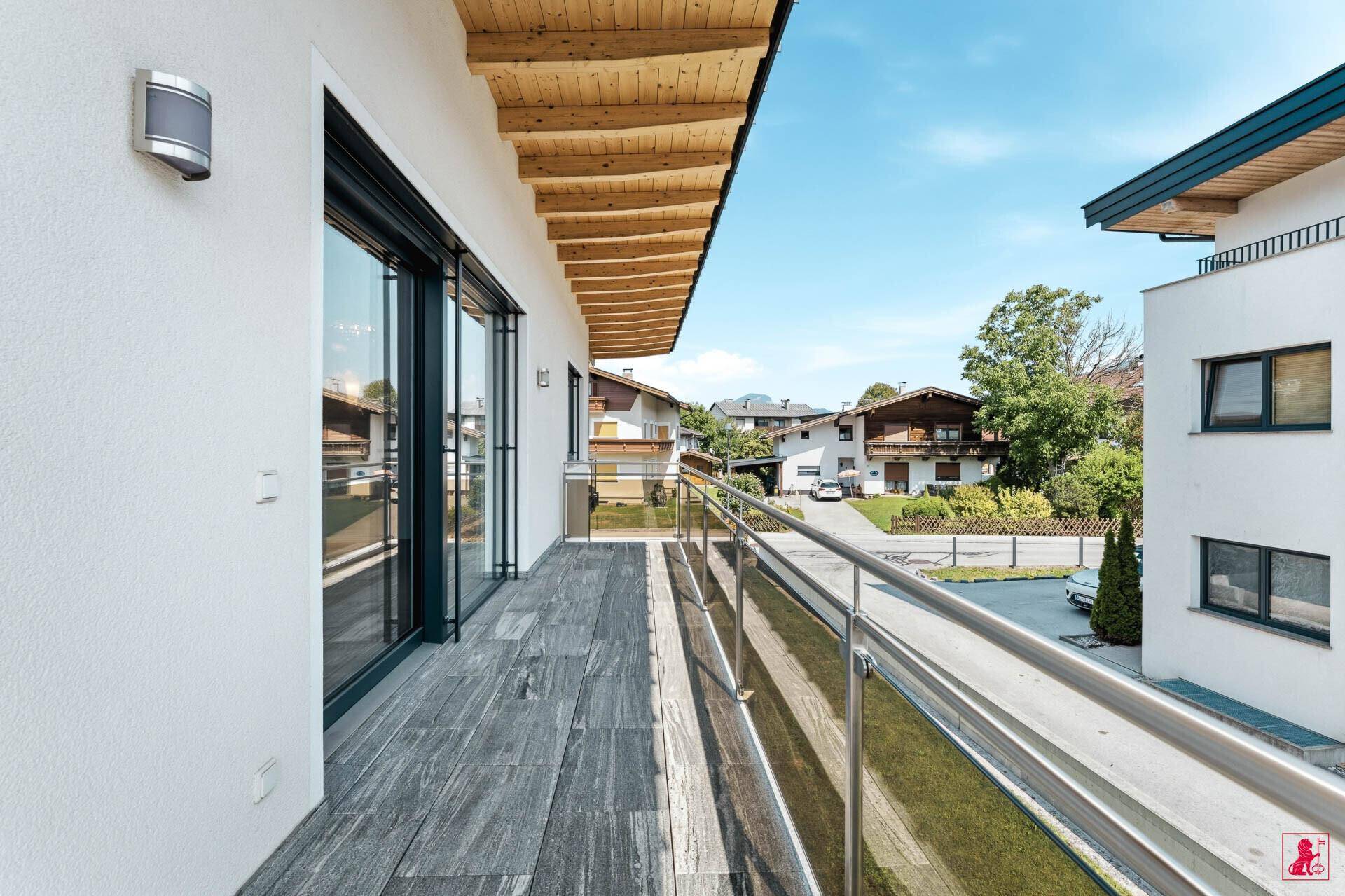 Langer Balkon mit Holzfliesen, Glasgeländer und Blick auf die Nachbarschaft.