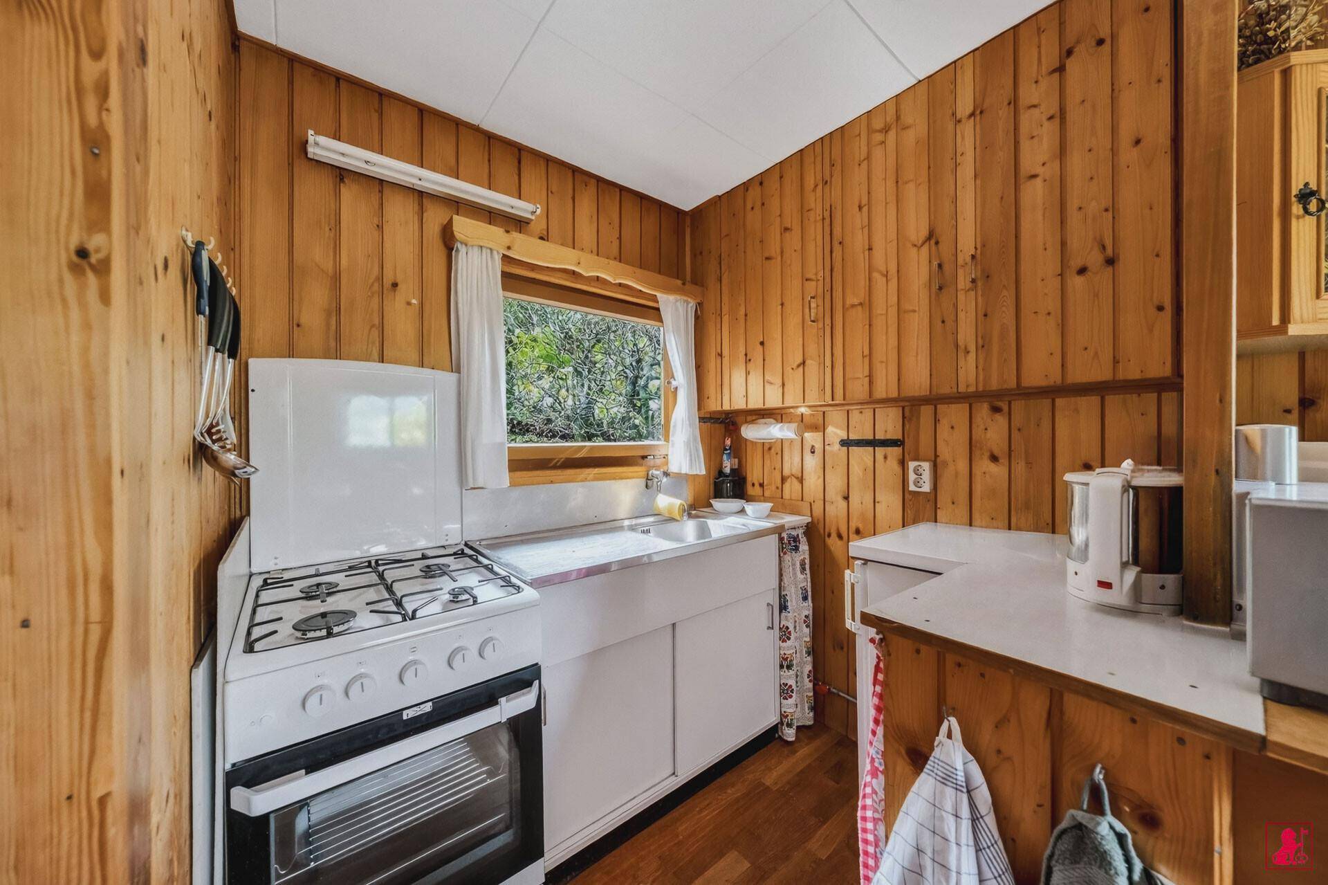 Kleine Küche mit Holzvertäfelung, Gasherd und Spülbecken am Fenster.