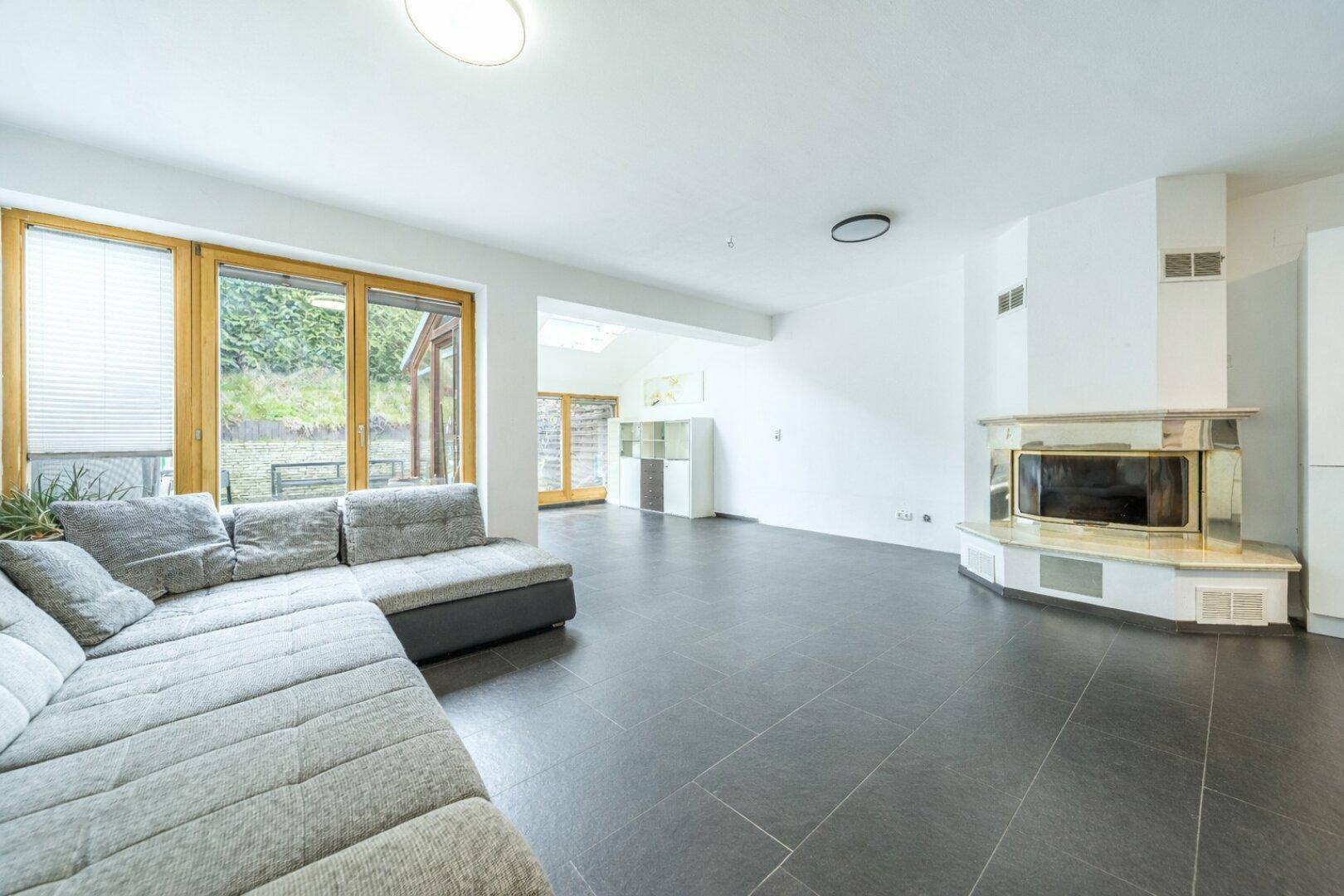 Geräumiges Wohnzimmer mit Kamin, großen Fenstern und dunklem Fliesenboden.