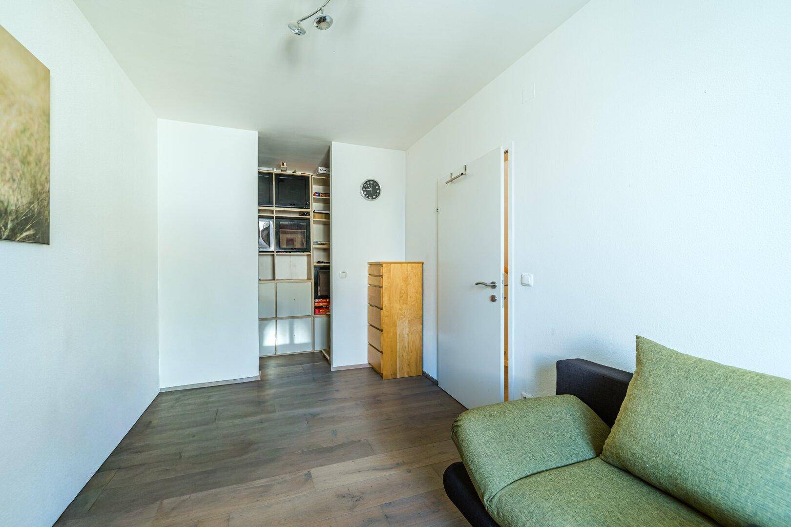 Kleiner Wohnbereich mit Sofa, Holzboden und Blick in einen Abstellraum.