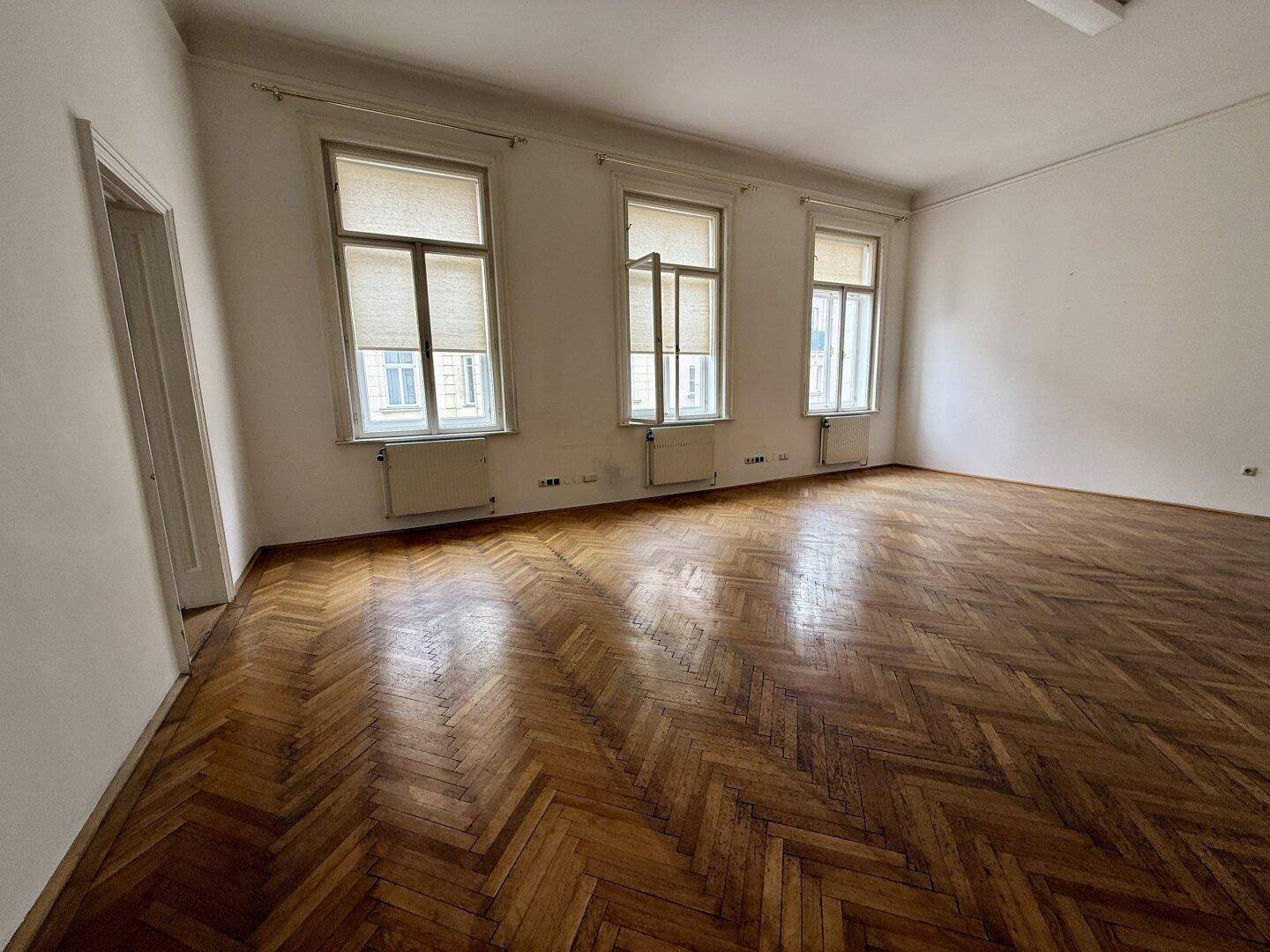 Großer Büroraum mit Fischgrätparkett und drei hohen Fenstern, die viel Tageslicht hereinlassen.