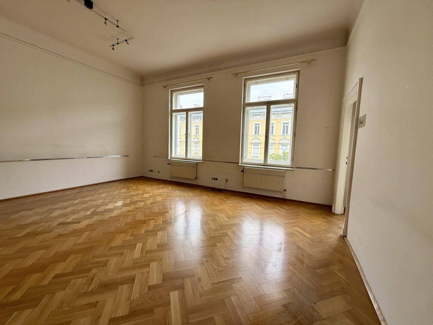 Heller Büroraum mit Fischgrätparkett und zwei großen Fenstern, die einen Blick nach draußen bieten.