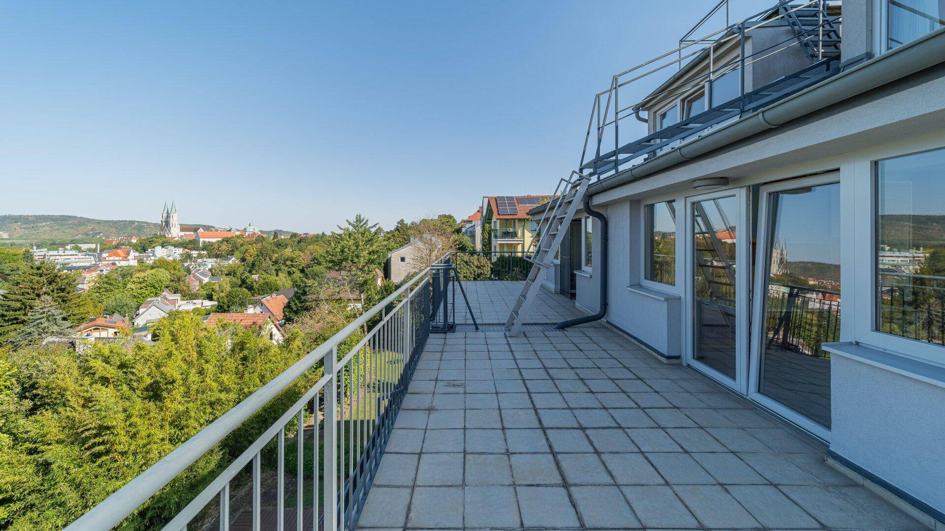 Geräumige Fliesenterrasse mit Metallgeländer und weitem Blick über die Stadt und die umliegende grüne Landschaft.