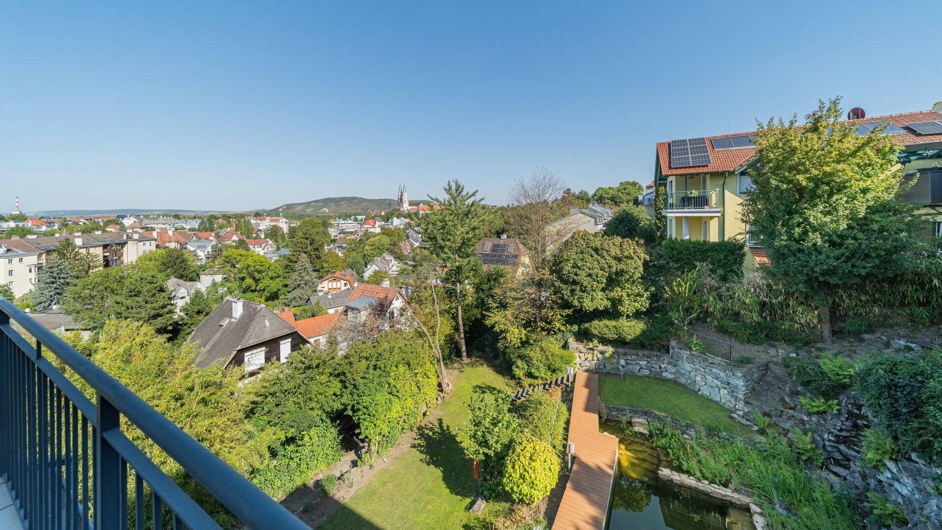 Weitläufiger Ausblick von einem Balkon über eine grüne Stadtlandschaft mit Häusern und einem Teich im Vordergrund.