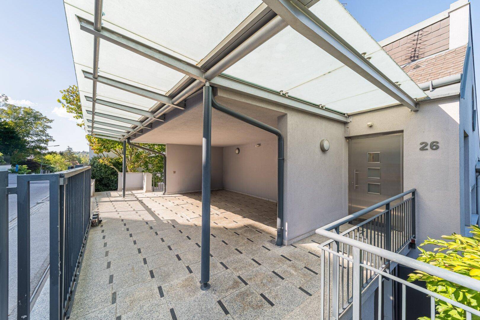 Eingangsbereich des Gebäudes mit überdachtem Carport, gefliestem Weg und moderner Haustür.