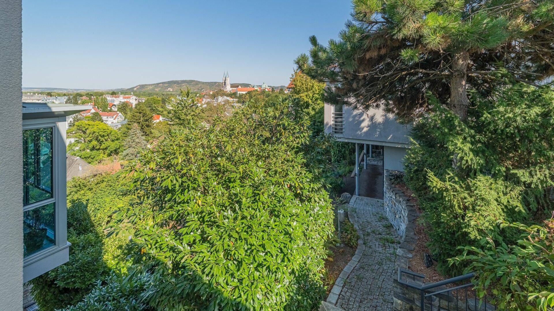 Blick auf einen gepflasterten Weg, der zu einem Eingang führt, umgeben von üppigem Grün und Stadtpanorama.