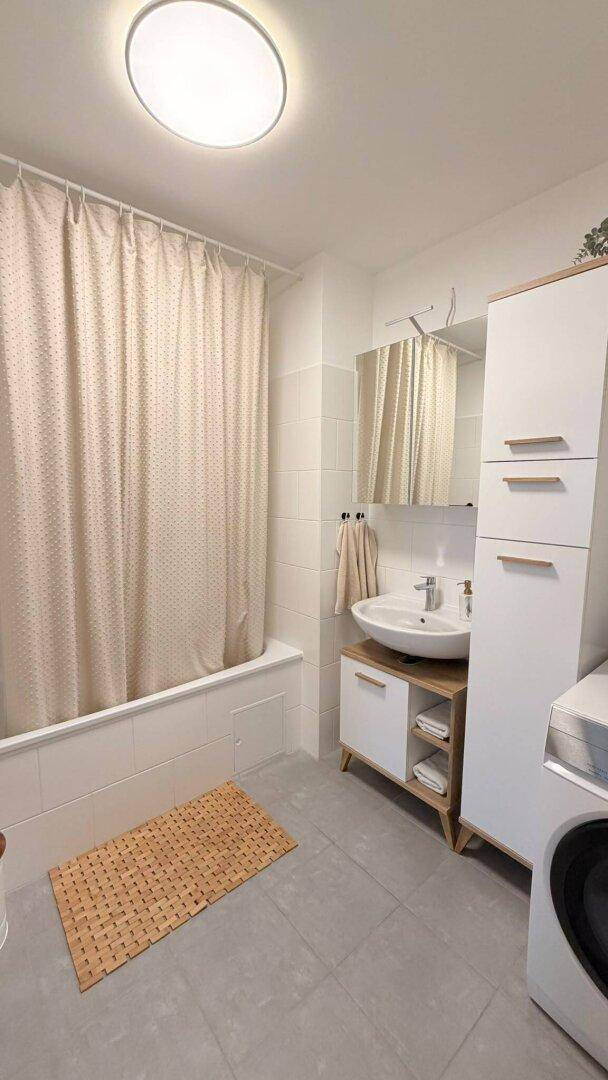 Badezimmer mit Badewanne, Duschvorhang, Einzelwaschbecken und Waschmaschine, auf grauen Fliesen.
