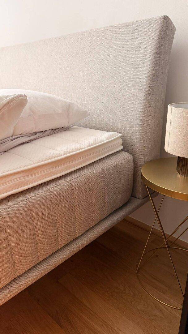 Nahaufnahme eines Bettes mit beigem Kopfteil und Matratze, auf einem Holzboden.