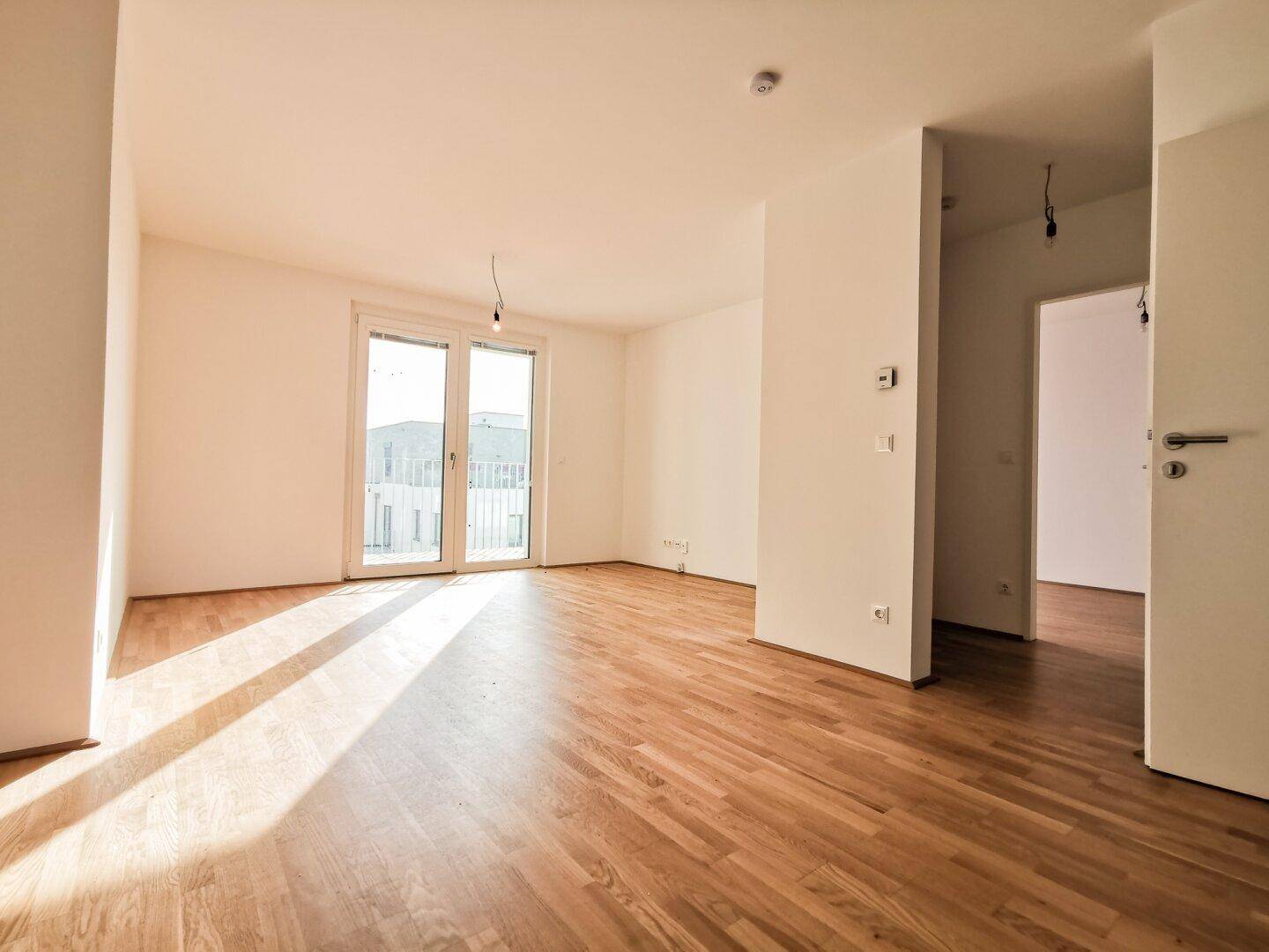 Heller, leerer Wohnraum mit Parkettboden und Zugang zu einem Balkon, durchflutet von natürlichem Licht.