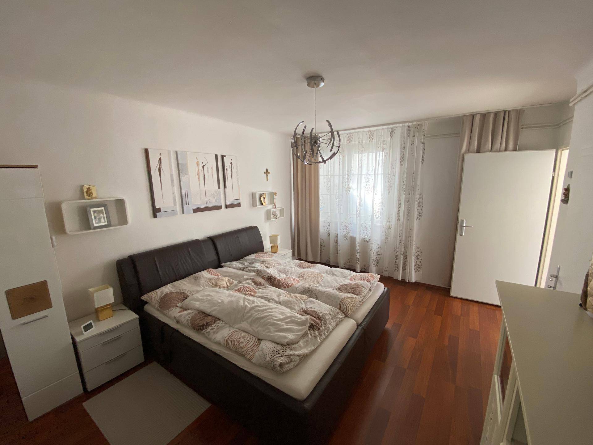 Helles Schlafzimmer mit Doppelbett, Fenster und Vorhängen auf Holzboden.