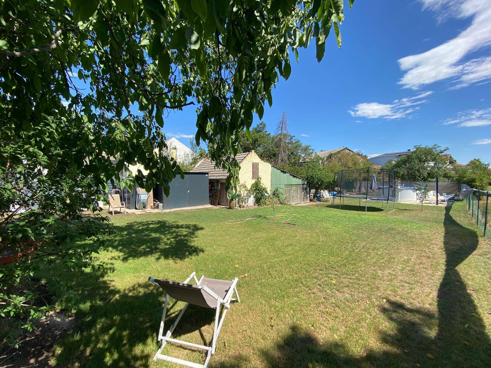 Großer Garten mit Trampolin, Schuppen und altem Baumbestand unter blauem Himmel.