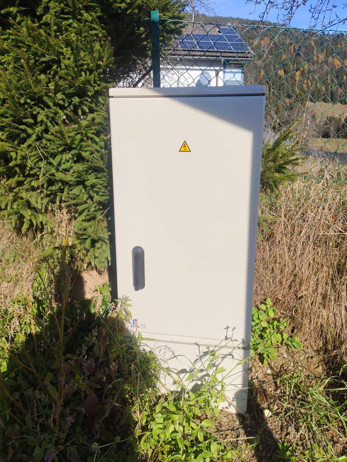 Grauer Stromverteilerkasten am Straßenrand, umgeben von Vegetation und einem Zaun.