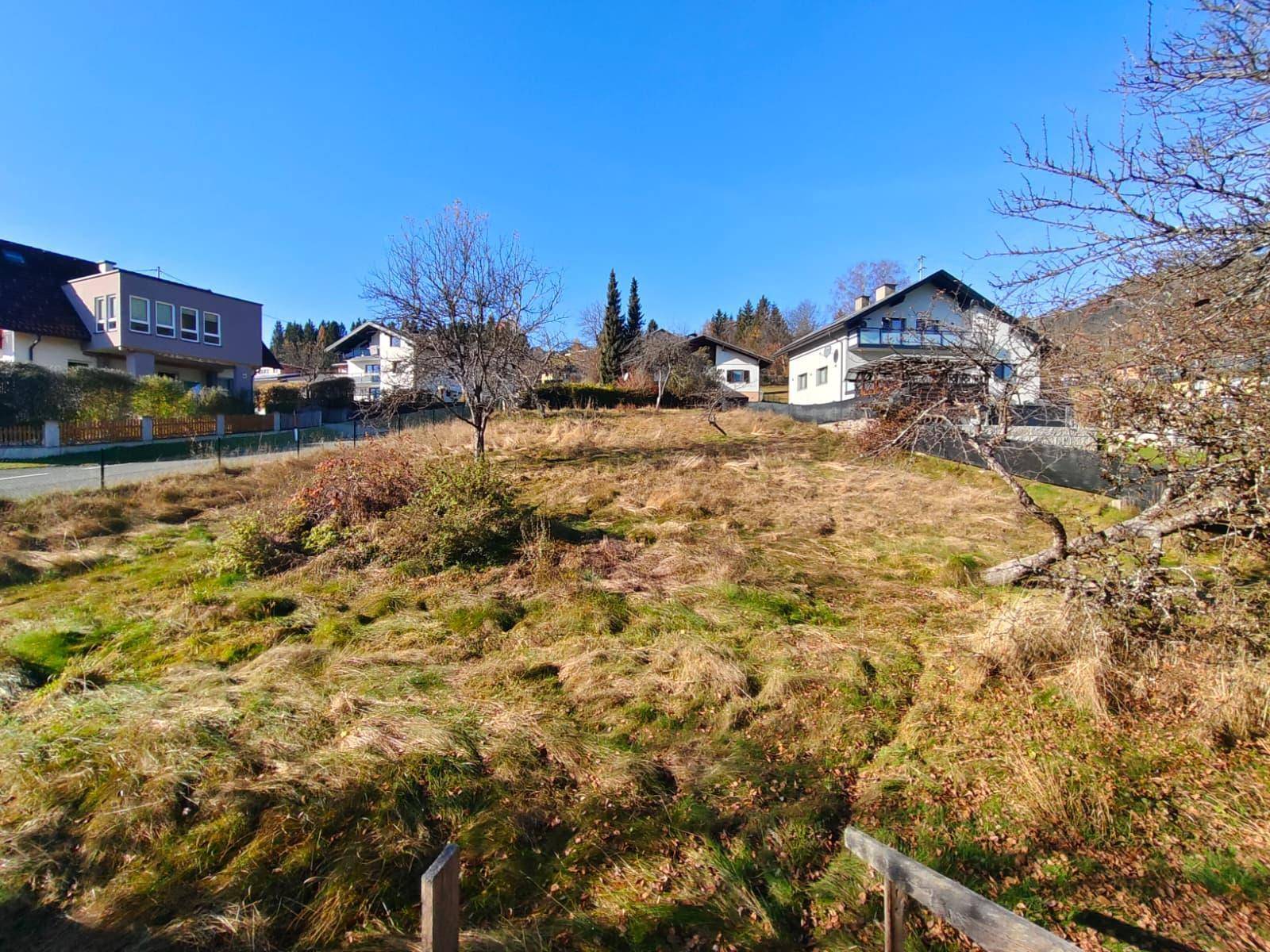Sonnenbeschienenes Grundstück mit trockenem Gras und Häusern im Hintergrund, umgeben von Natur.