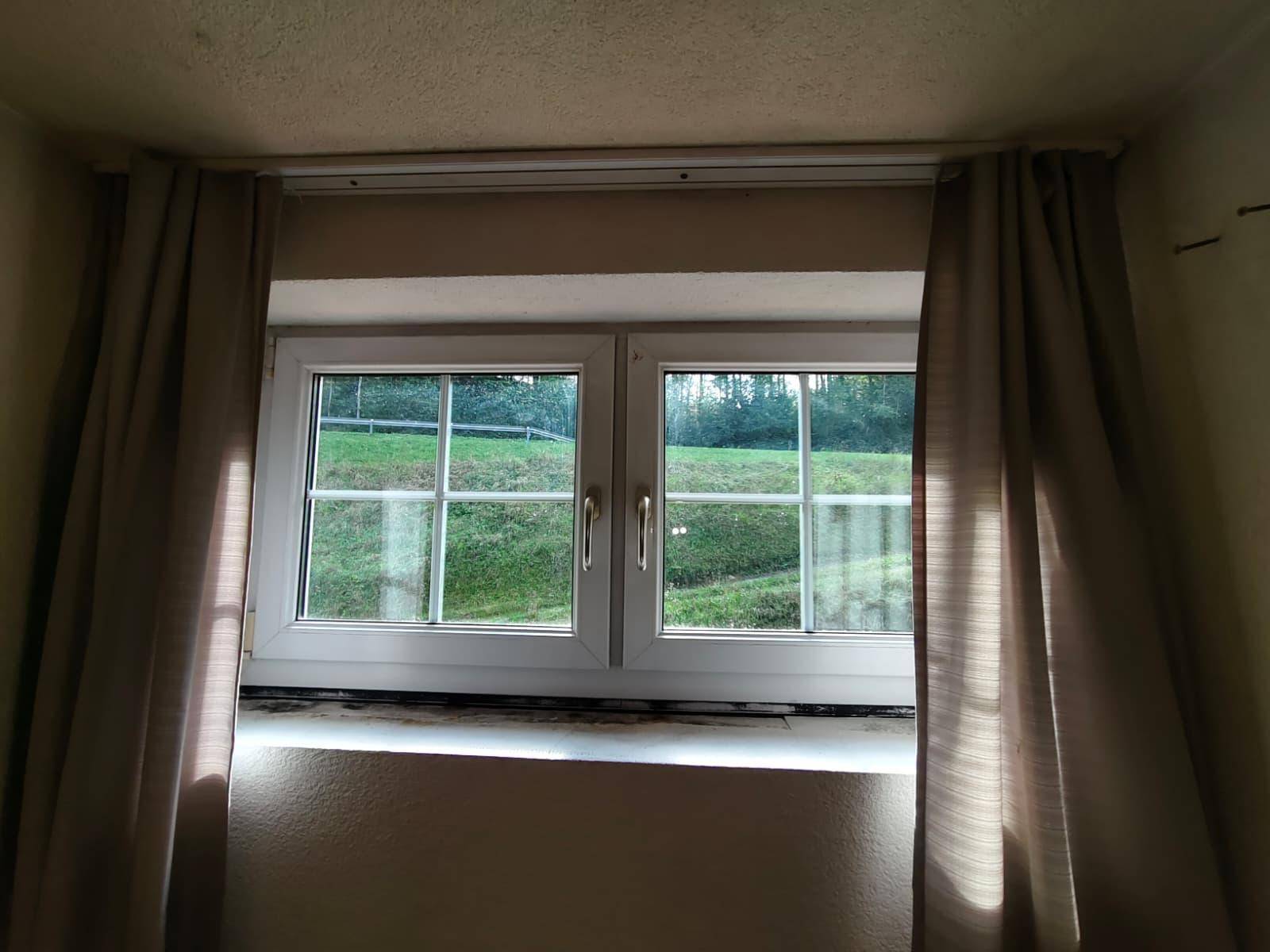 Ein Fenster mit Vorhängen bietet einen Ausblick auf eine grüne Wiese und die umliegende Natur.
