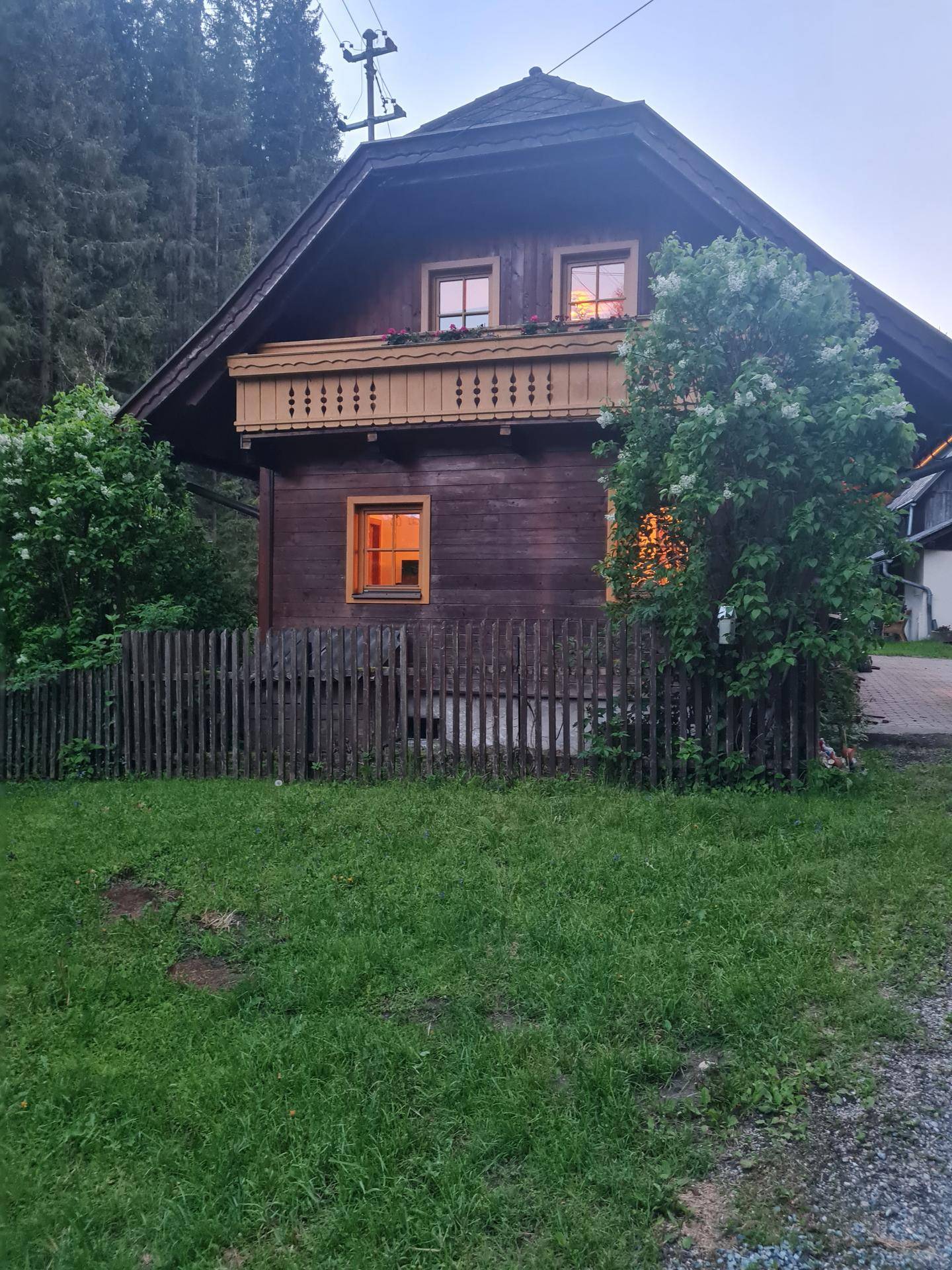 Traditionelles Holzhaus mit Balkon, umzäunt von einem Holzzaun und grünem Rasen in der Abenddämmerung.