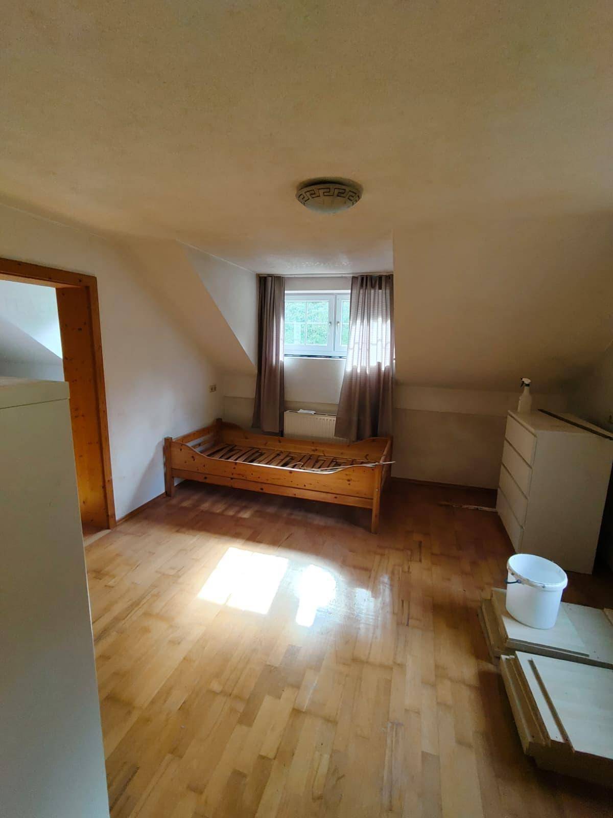 Dachzimmer mit Einzelbettgestell, Kommode und einem Fenster mit Vorhängen, unter schrägen Decken.