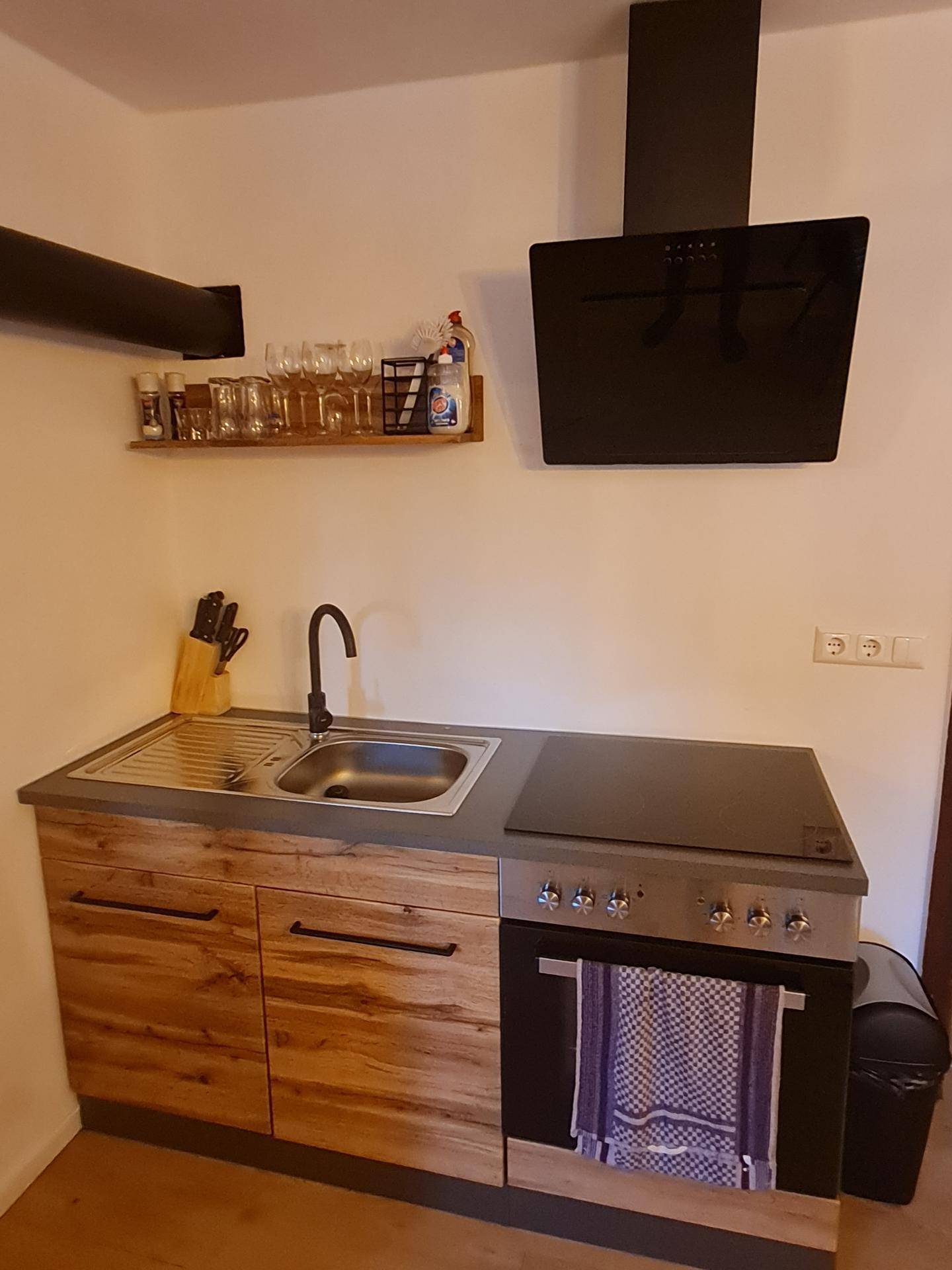 Kleine Küchenzeile mit Holzschränken, Spüle, Kochfeld, Backofen und schwarzer Dunstabzugshaube.