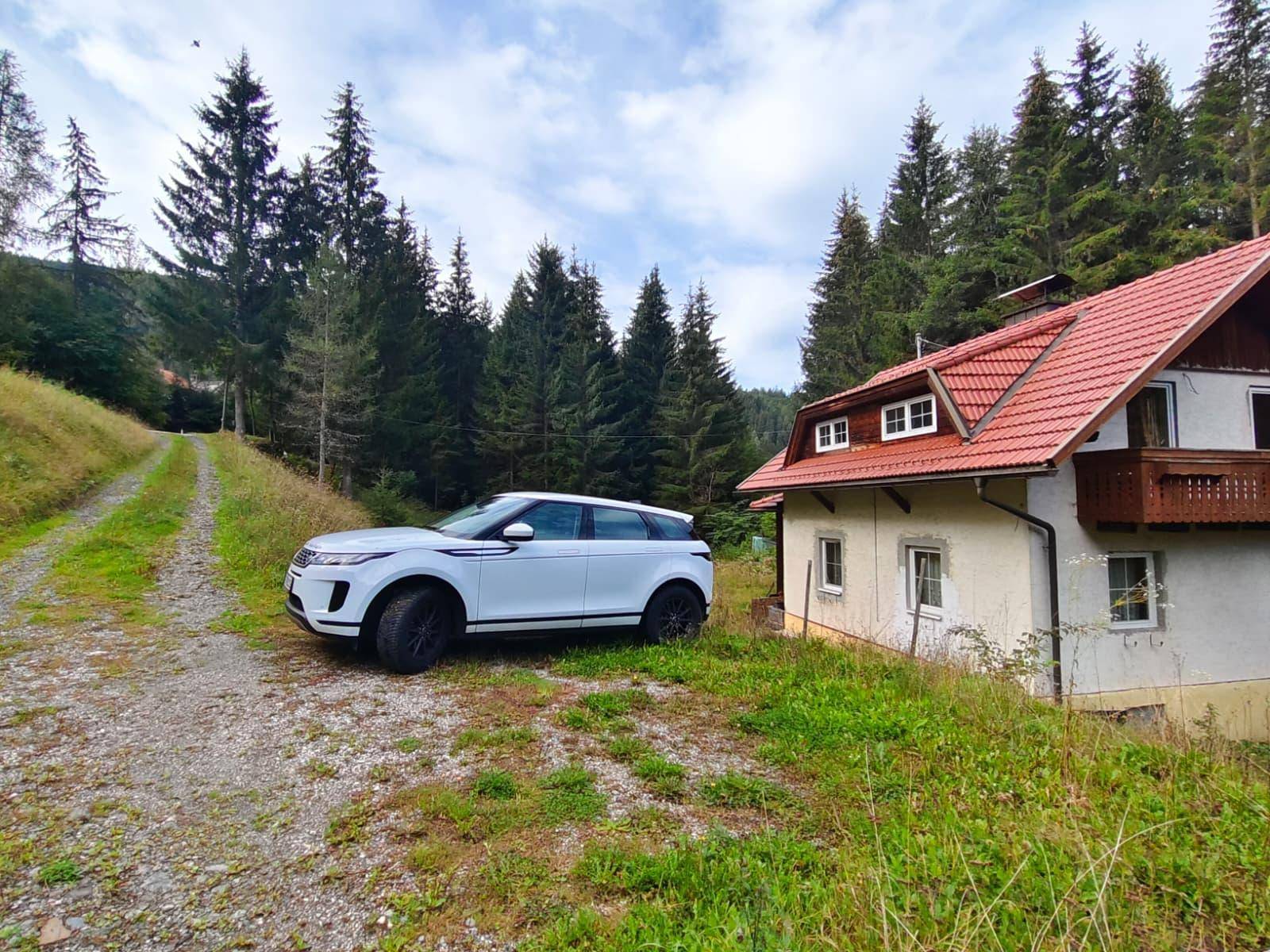 Seitenansicht des Hauses mit einem weißen Auto auf einem Schotterweg, umgeben von Wald.