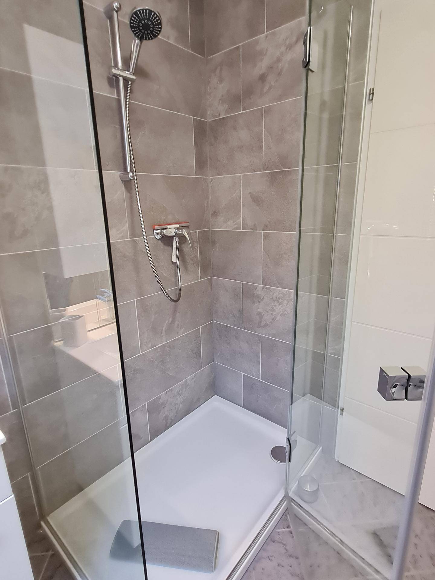 Moderne Duschkabine mit Glasabtrennung und grauen Wandfliesen in einem hellen Badezimmer.