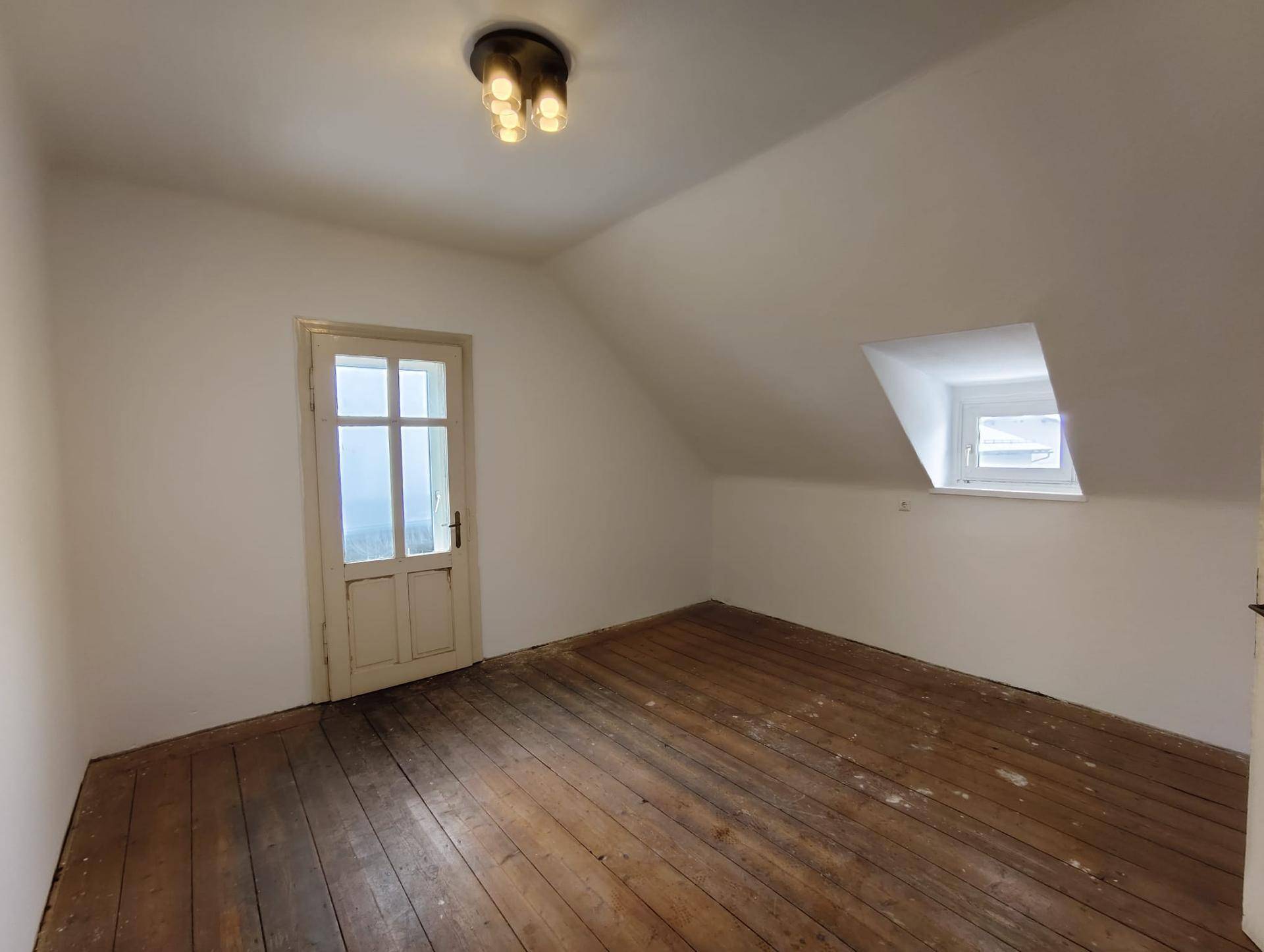 Geräumiges Dachzimmer mit schrägen Wänden, altem Holzboden und einem Fenster für natürliches Licht.