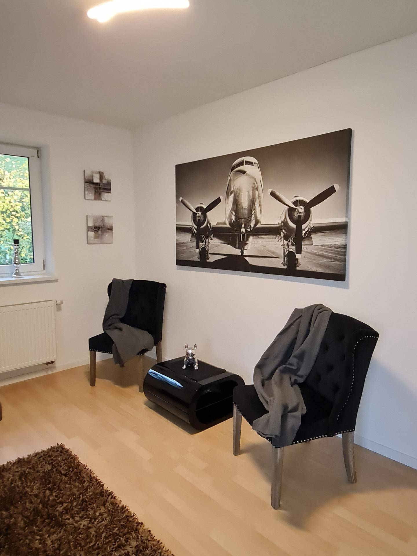 Helles Wohnzimmer mit zwei schwarzen Sesseln, Couchtisch und großem Flugzeugbild an der Wand.