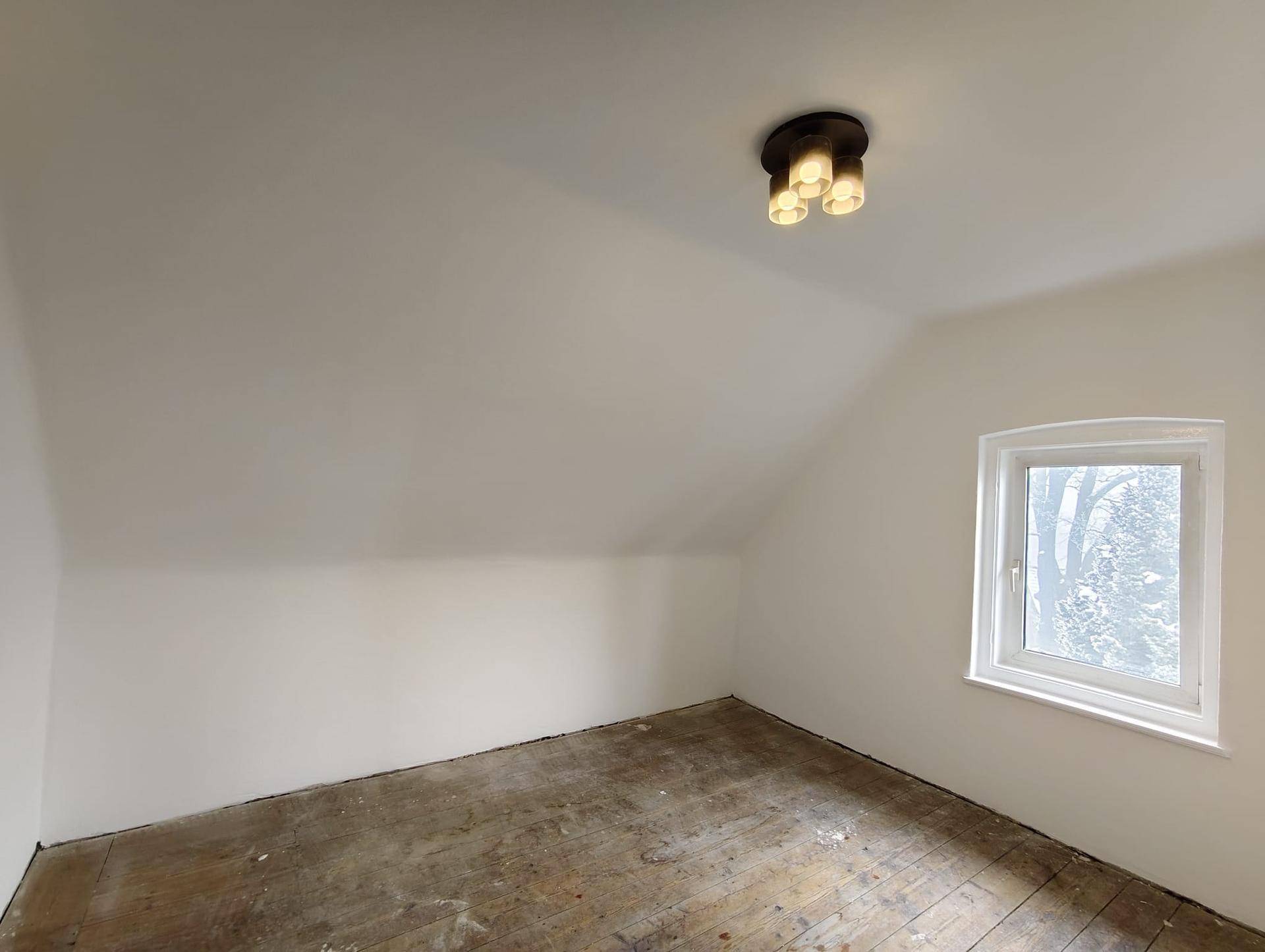 Leeres Zimmer im Dachgeschoss mit schrägen Wänden, altem Holzboden und kleinem Fenster.