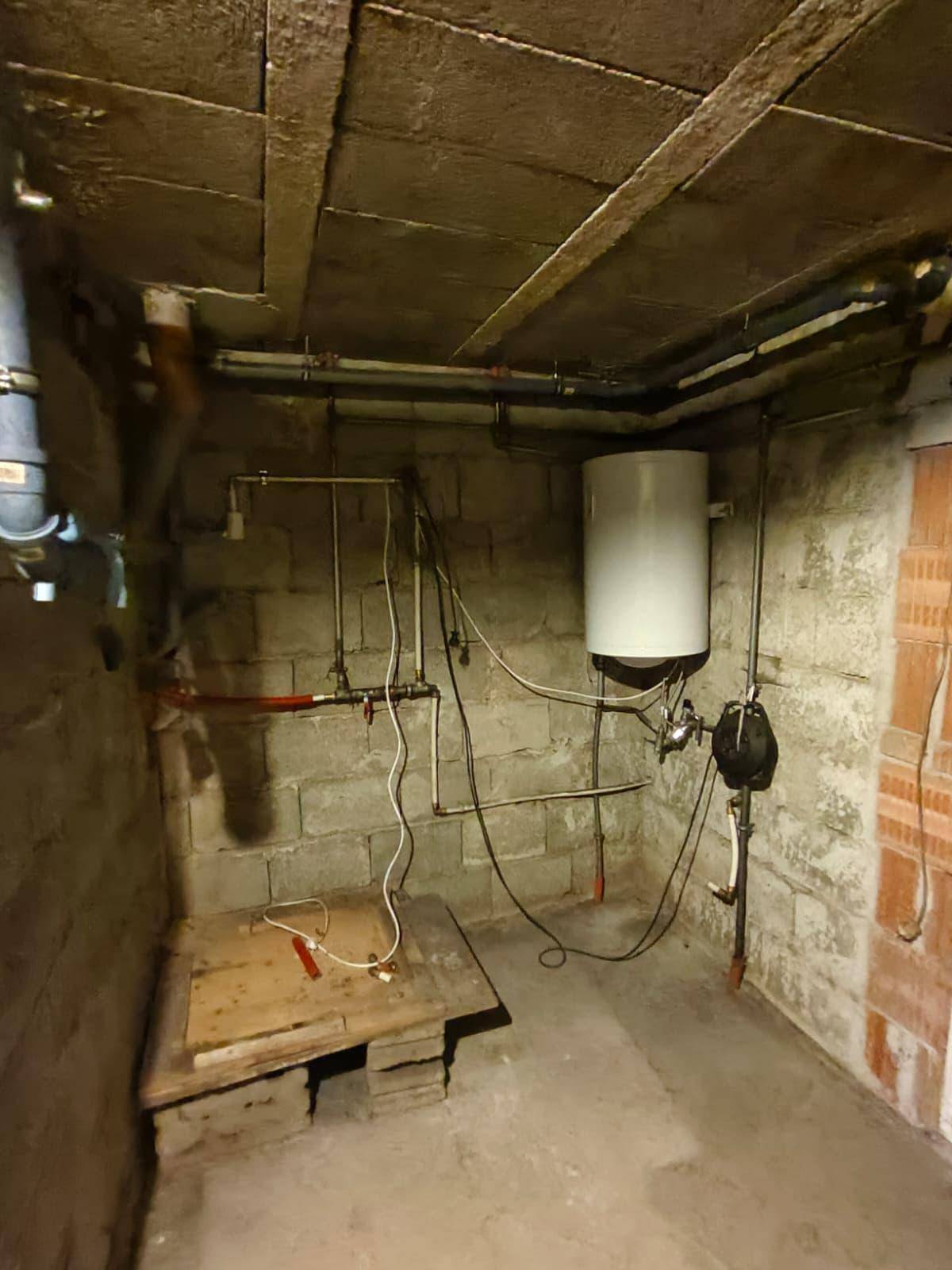 Teil eines Kellerraums mit sichtbaren Rohren, einem Warmwasserbereiter und unverputzten Betonwänden.