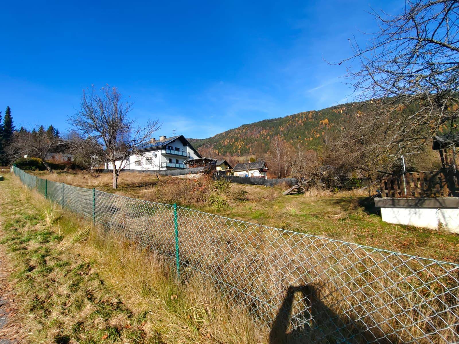Großes Grundstück mit Maschendrahtzaun, einem Haus und bewaldeten Bergen im Hintergrund.