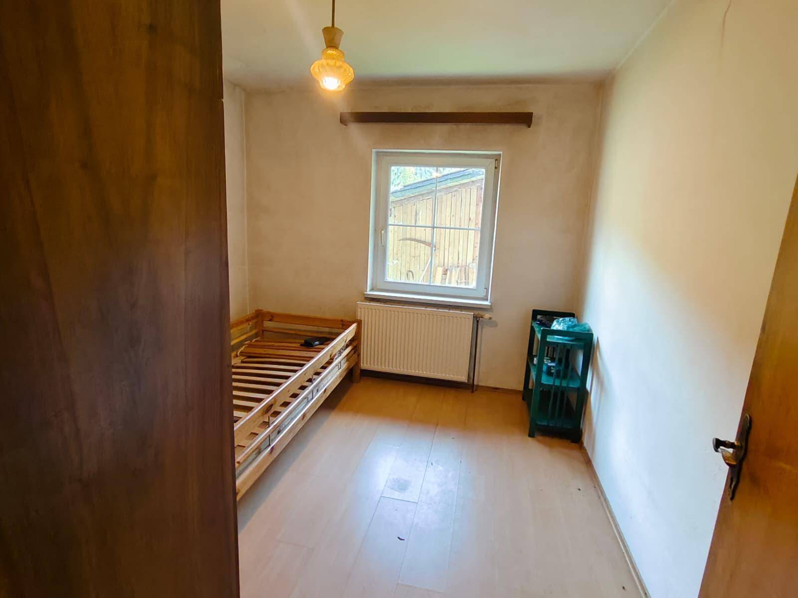 Kleines Schlafzimmer mit Einzelbettrahmen, hellem Boden und Fenster, das Tageslicht hereinlässt.