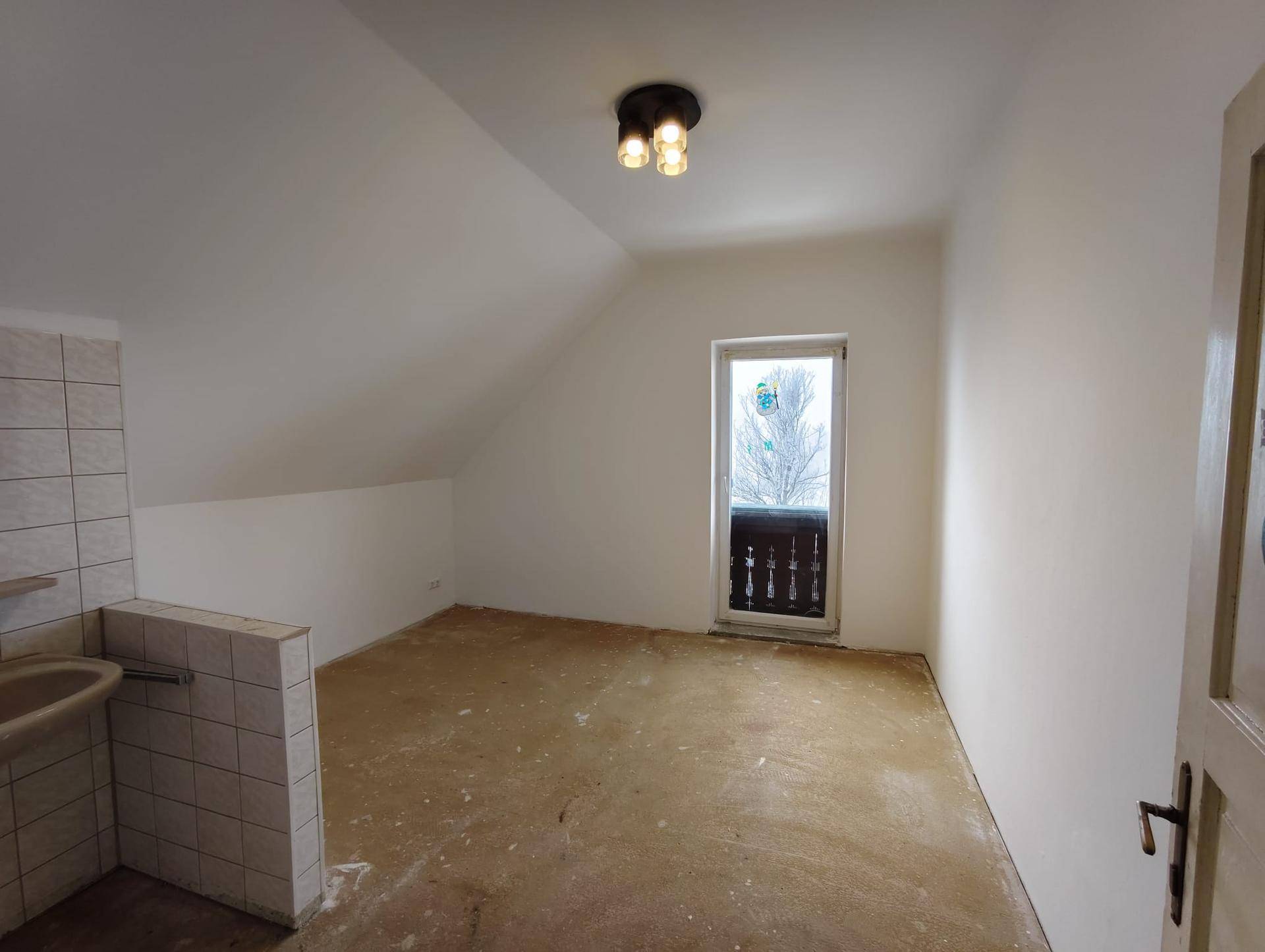 Dachzimmer mit schrägen Wänden, altem Boden, kleinem Waschbecken und Zugang zu einem französischen Balkon.