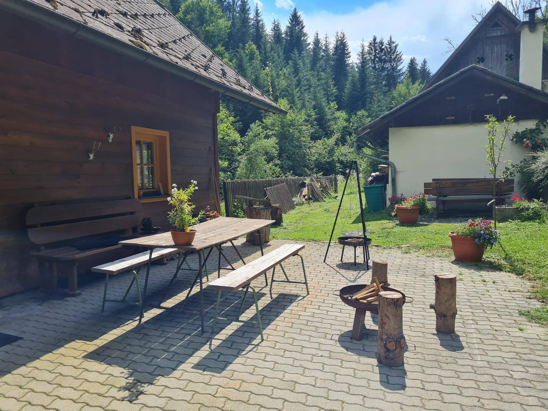 Gepflasterte Terrasse mit langem Holztisch und Bänken, Feuerstelle und Topfpflanzen, umgeben von Wald.