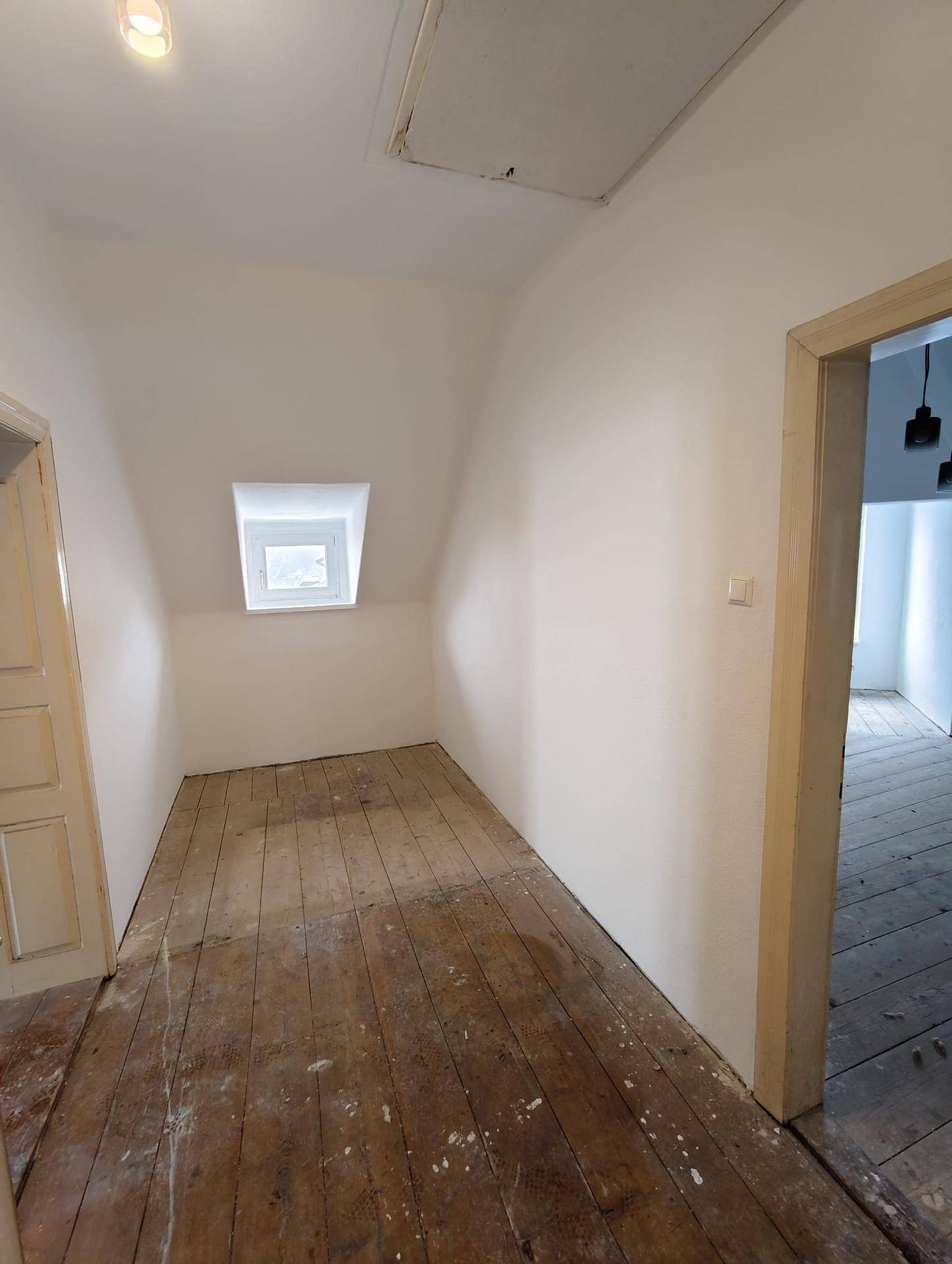 Dachzimmer mit schrägen Wänden, altem Holzboden und einem kleinen Fenster, offen zu einem weiteren Raum.