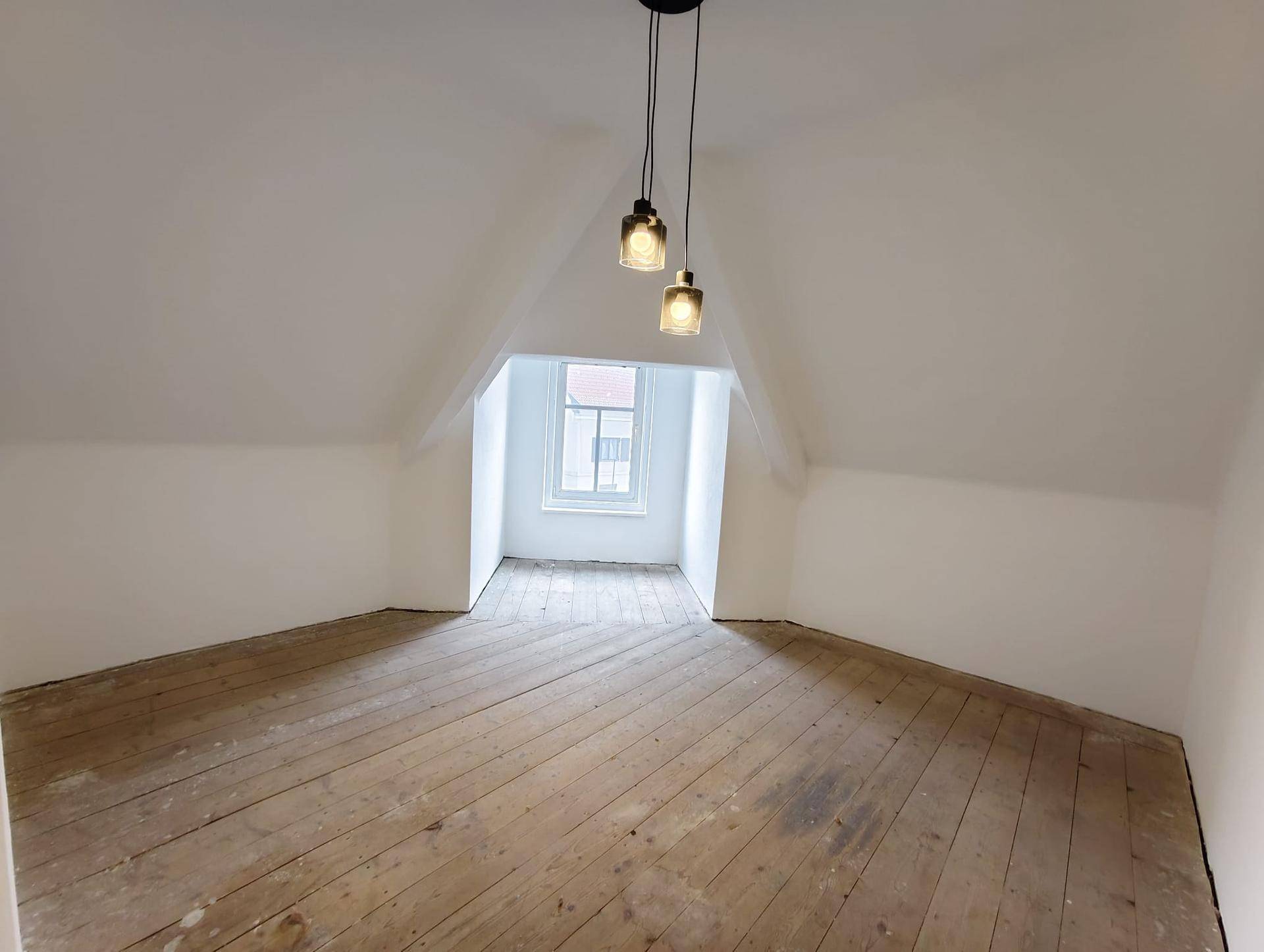 Helles Dachzimmer mit schrägen Wänden, altem Holzboden und einem kleinen Fenster.