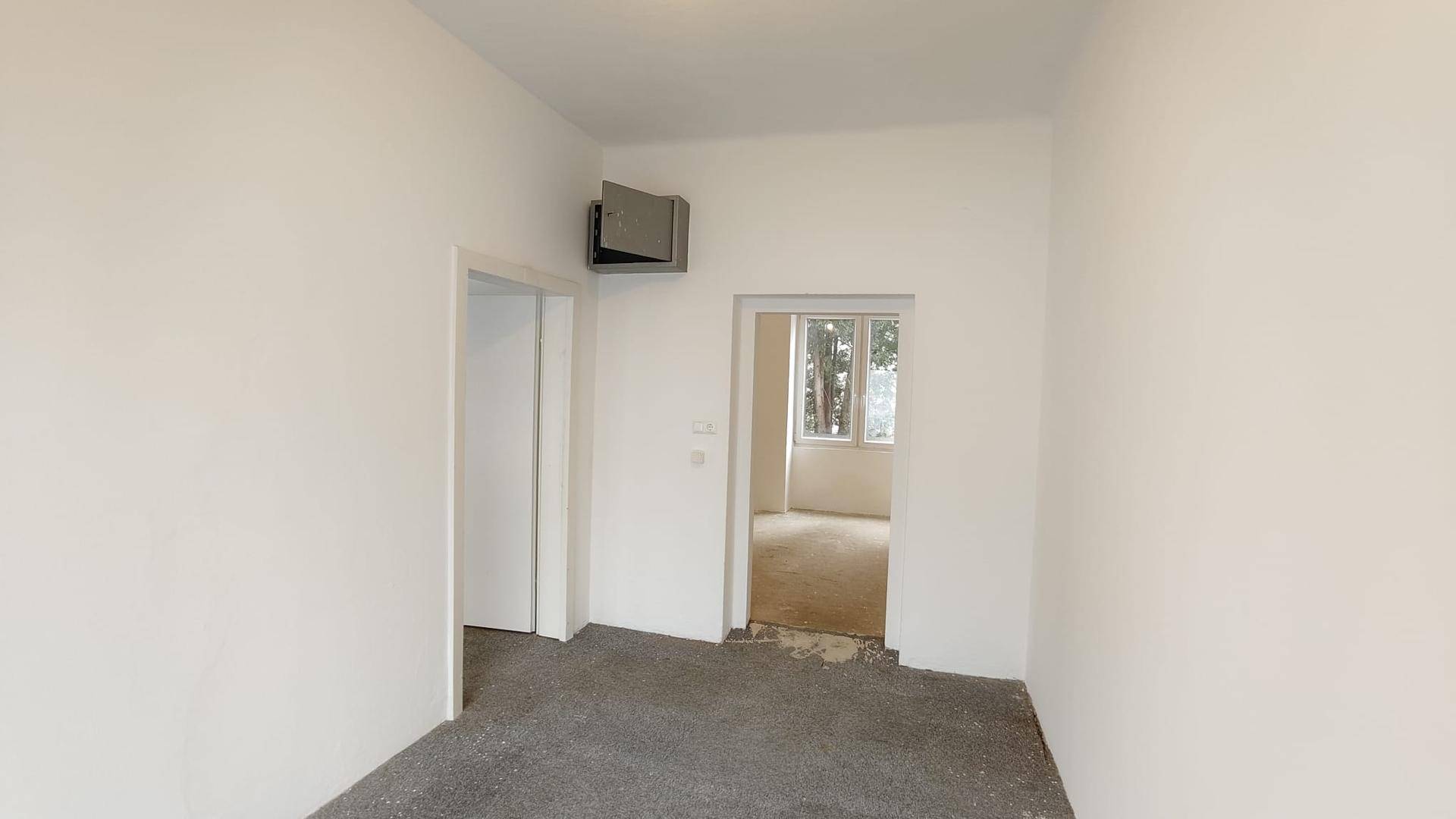 Heller Flur mit weißen Wänden, zwei Türen und Blick in einen angrenzenden Raum, mit Betonboden.