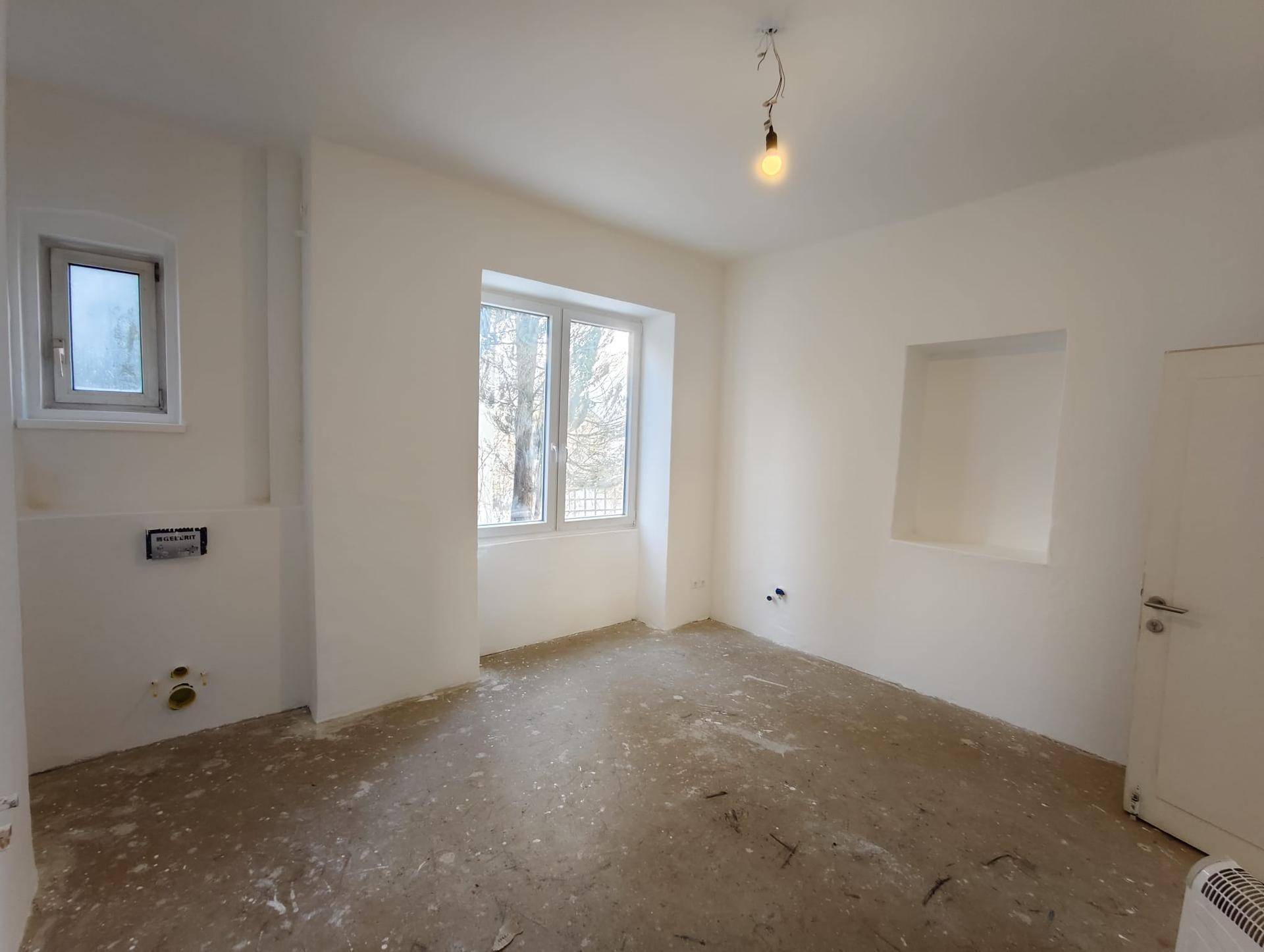 Renovierungsbedürftiger Raum mit einem Fenster, weißen Wänden und sichtbaren Wasseranschlüssen für ein Badezimmer.