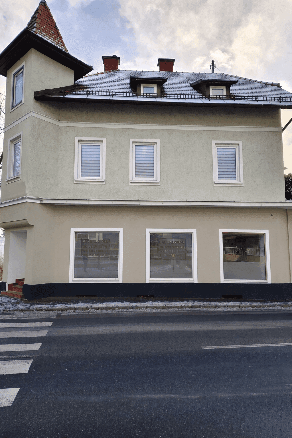 Seitenansicht des Gebäudes mit mehreren Fenstern, Rollläden und einem Zebrastreifen im Vordergrund an einem Wintertag.