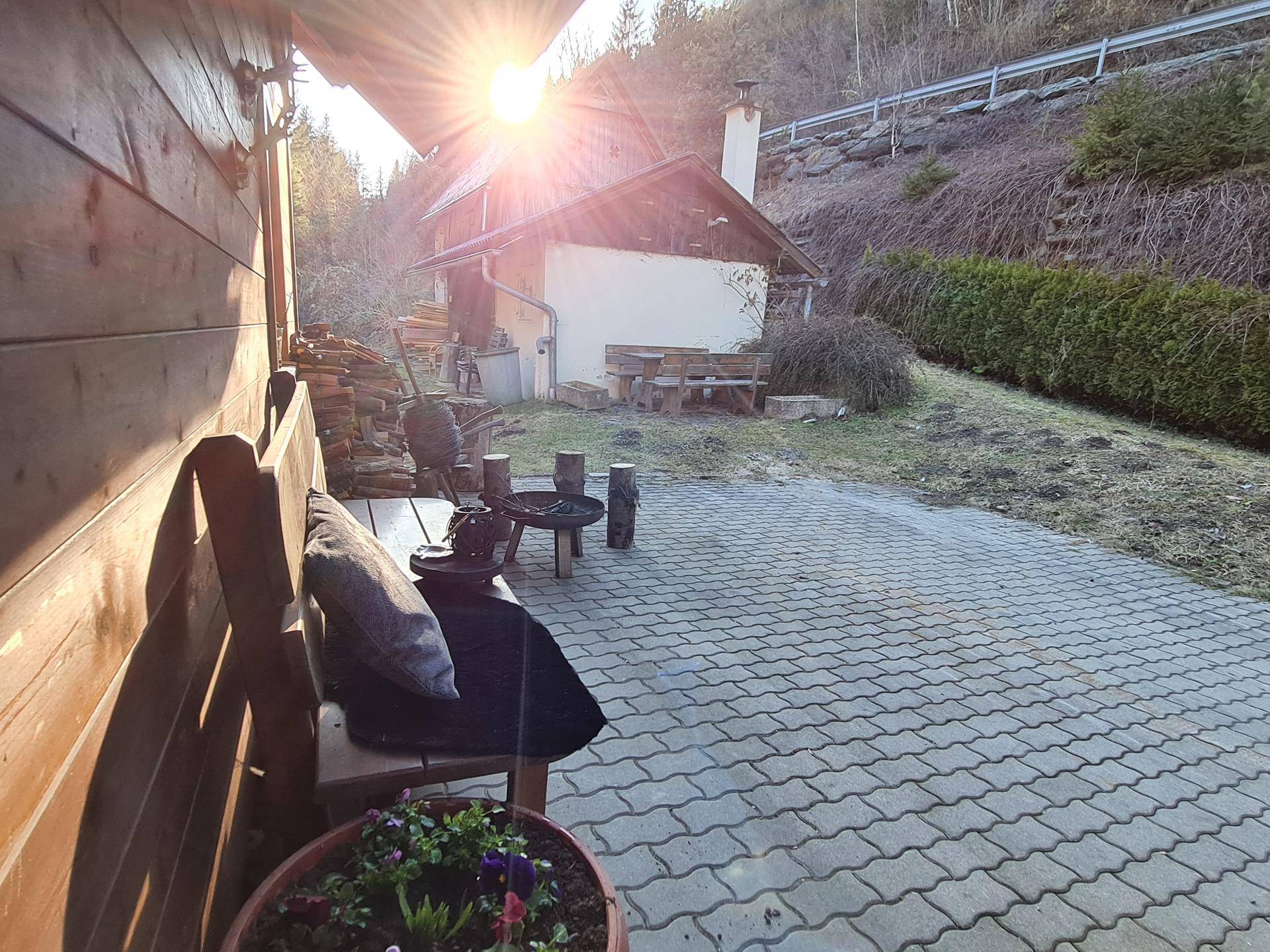 Gepflasterte Terrasse neben dem Holzhaus mit Sitzbank, Feuerstelle und Holzscheiten unter strahlendem Sonnenschein.
