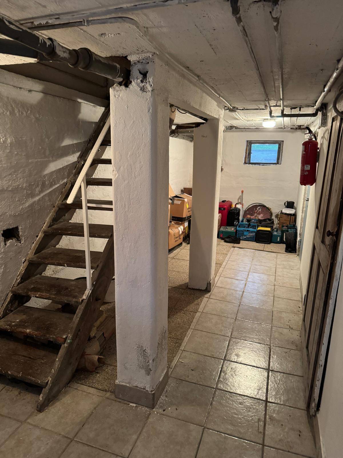 Kellerraum mit gefliestem Boden, Holztreppe und Lagerflächen für verschiedene Gegenstände.