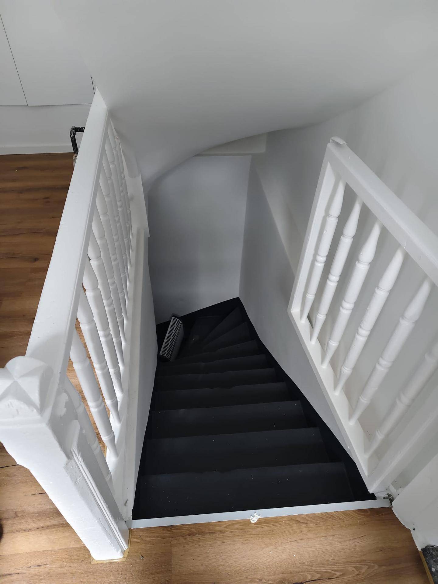 Blick von oben auf eine dunkle Treppe mit weißem Holzgeländer in einem hellen Treppenhaus.