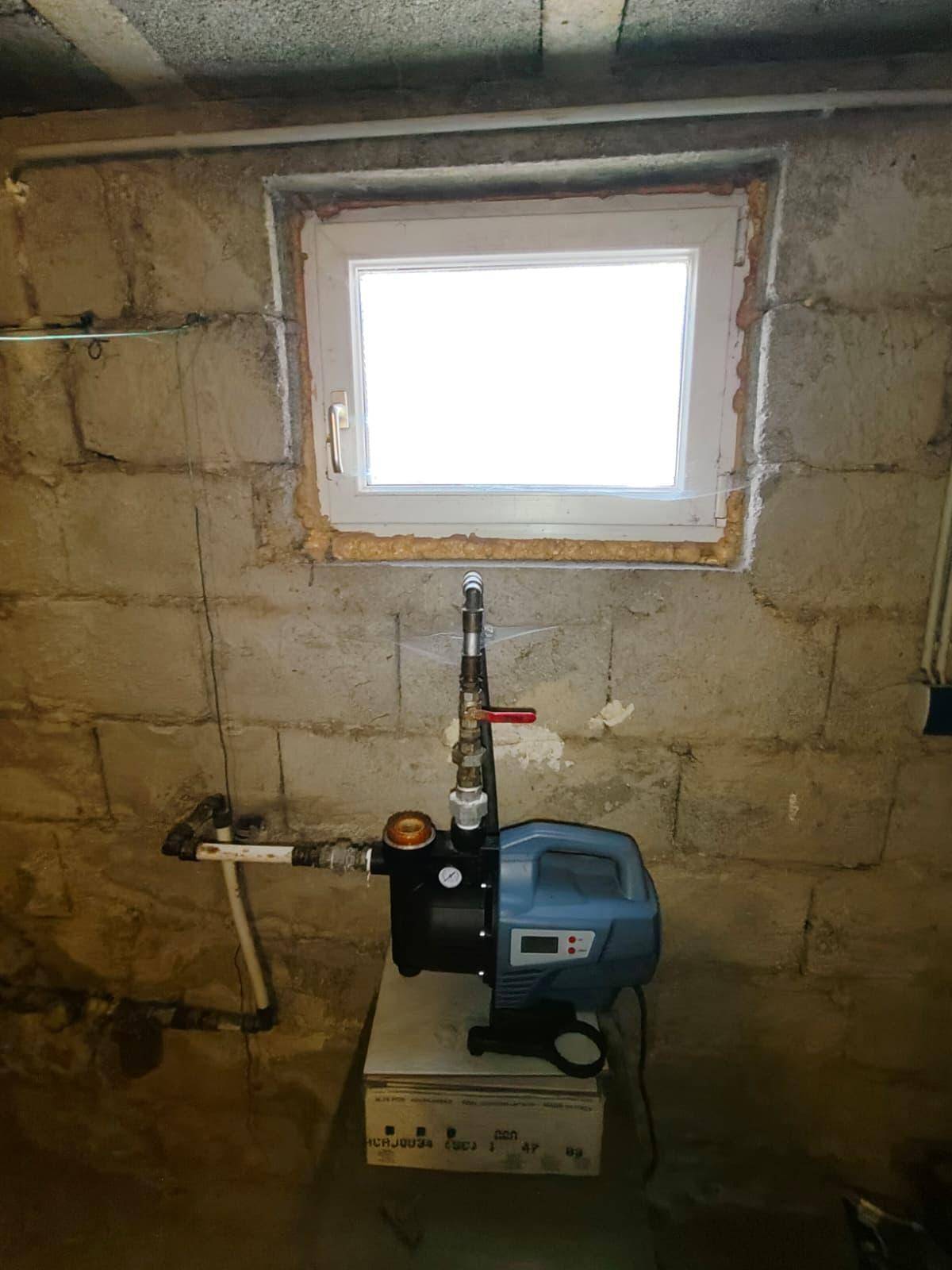 Nahaufnahme eines Fensters in einer Betonwand mit einer Wasserpumpe darunter, typisch für einen Kellerraum.