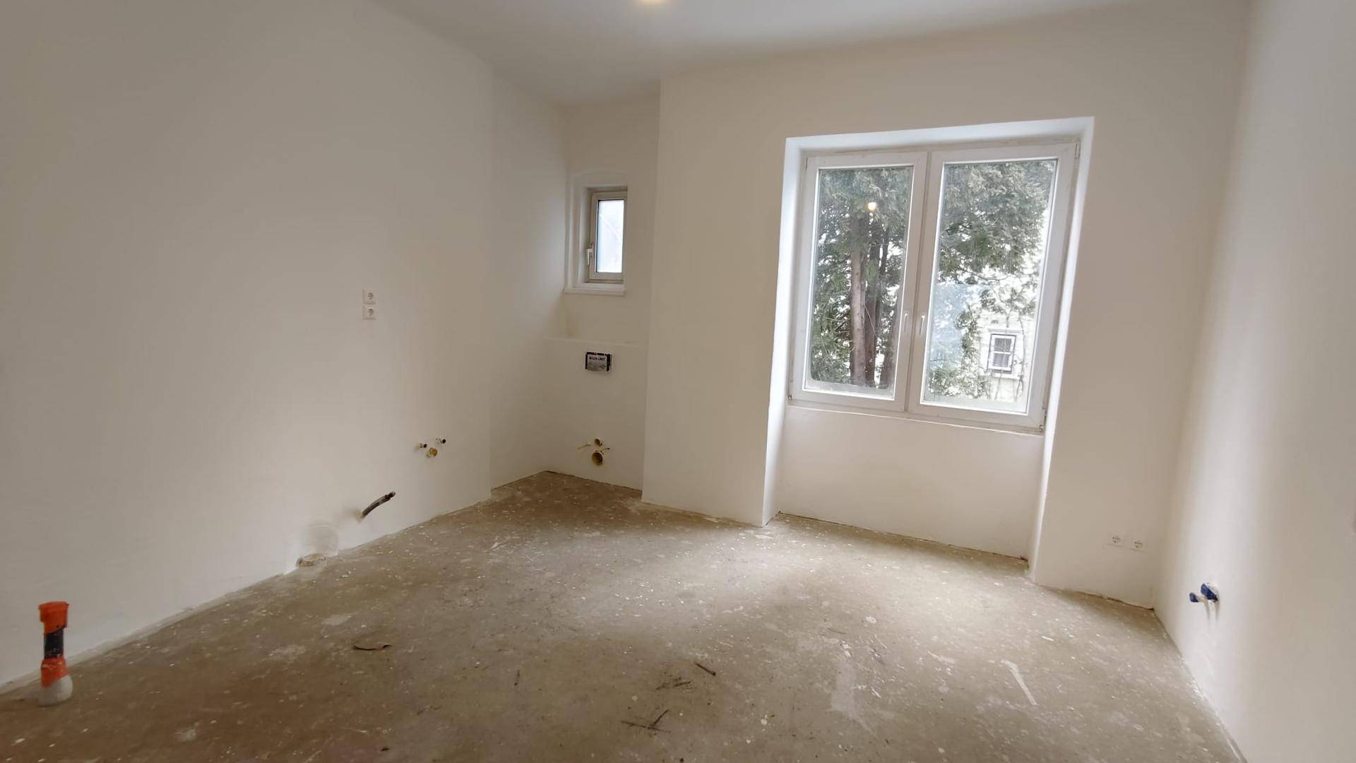 Renovierter Raum mit einem großen Fenster, weißen Wänden und sichtbaren Installationen für ein Badezimmer.