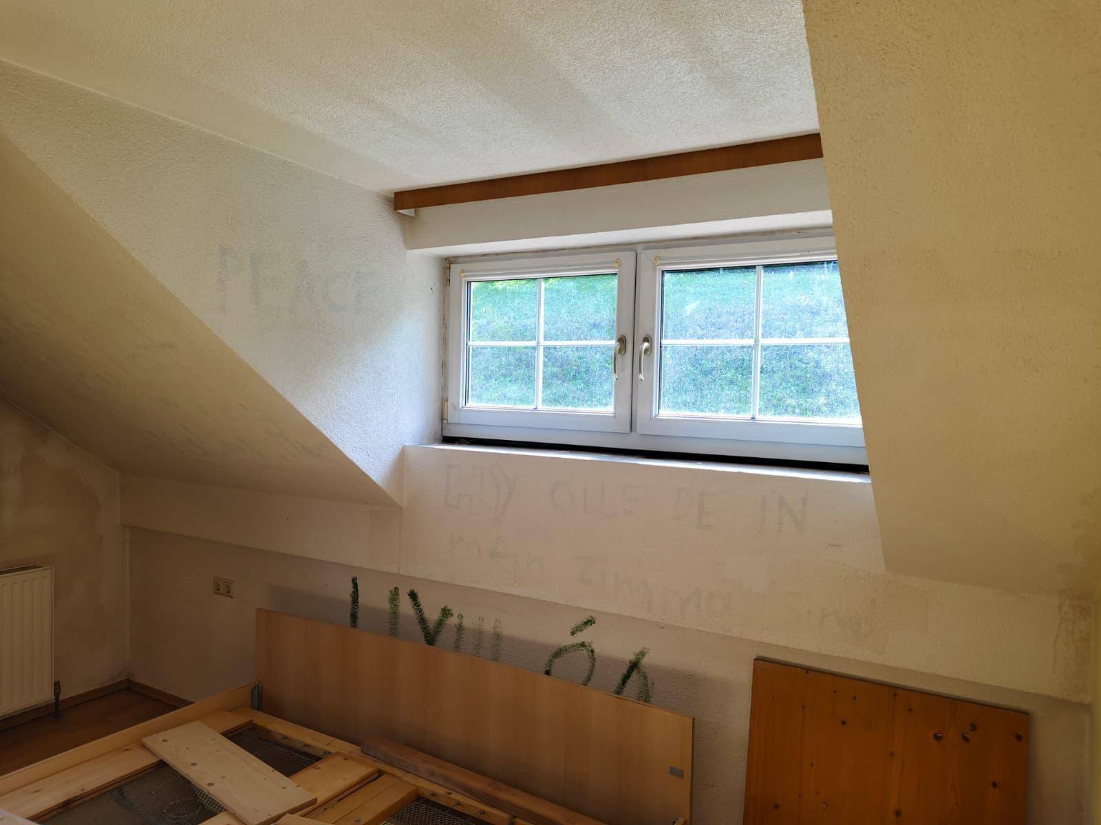 Dachzimmer mit schrägen Wänden, einem Fenster und einem Bettgestell, bereit zur Renovierung.