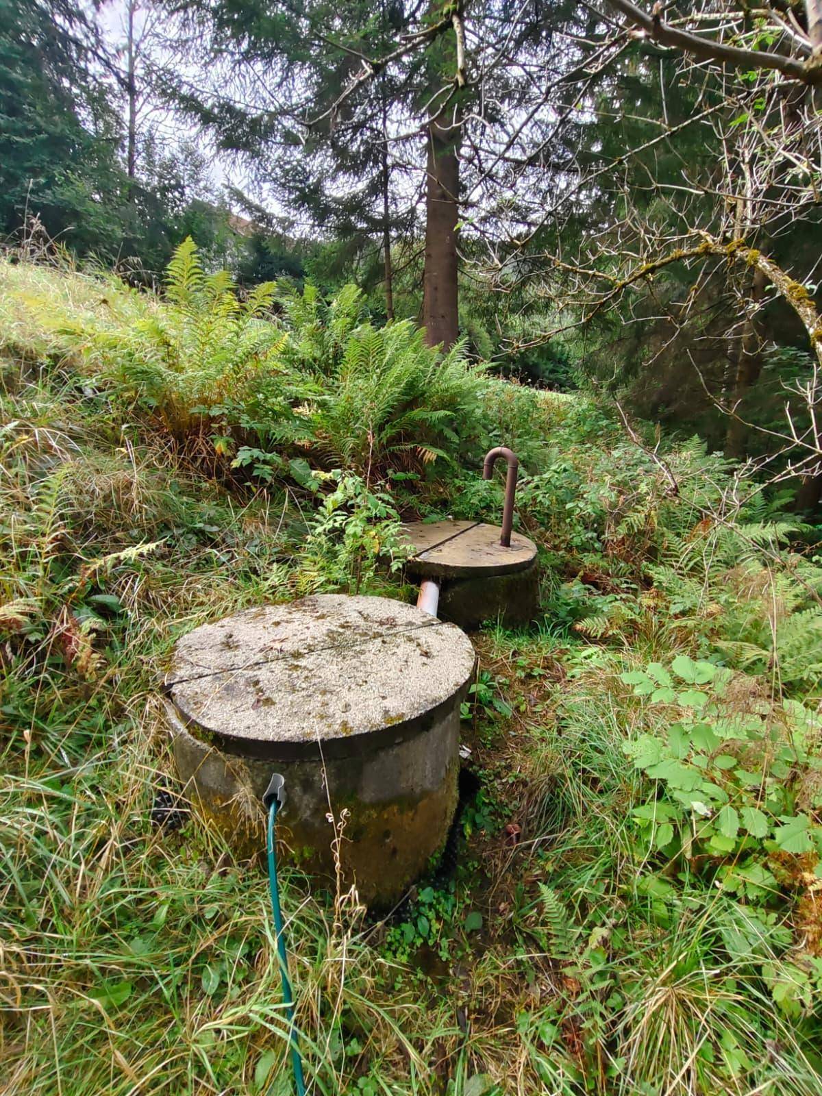Zwei Betonstrukturen in einem grasbewachsenen Bereich nahe Bäumen, möglicherweise für Wasser oder Abwasser.