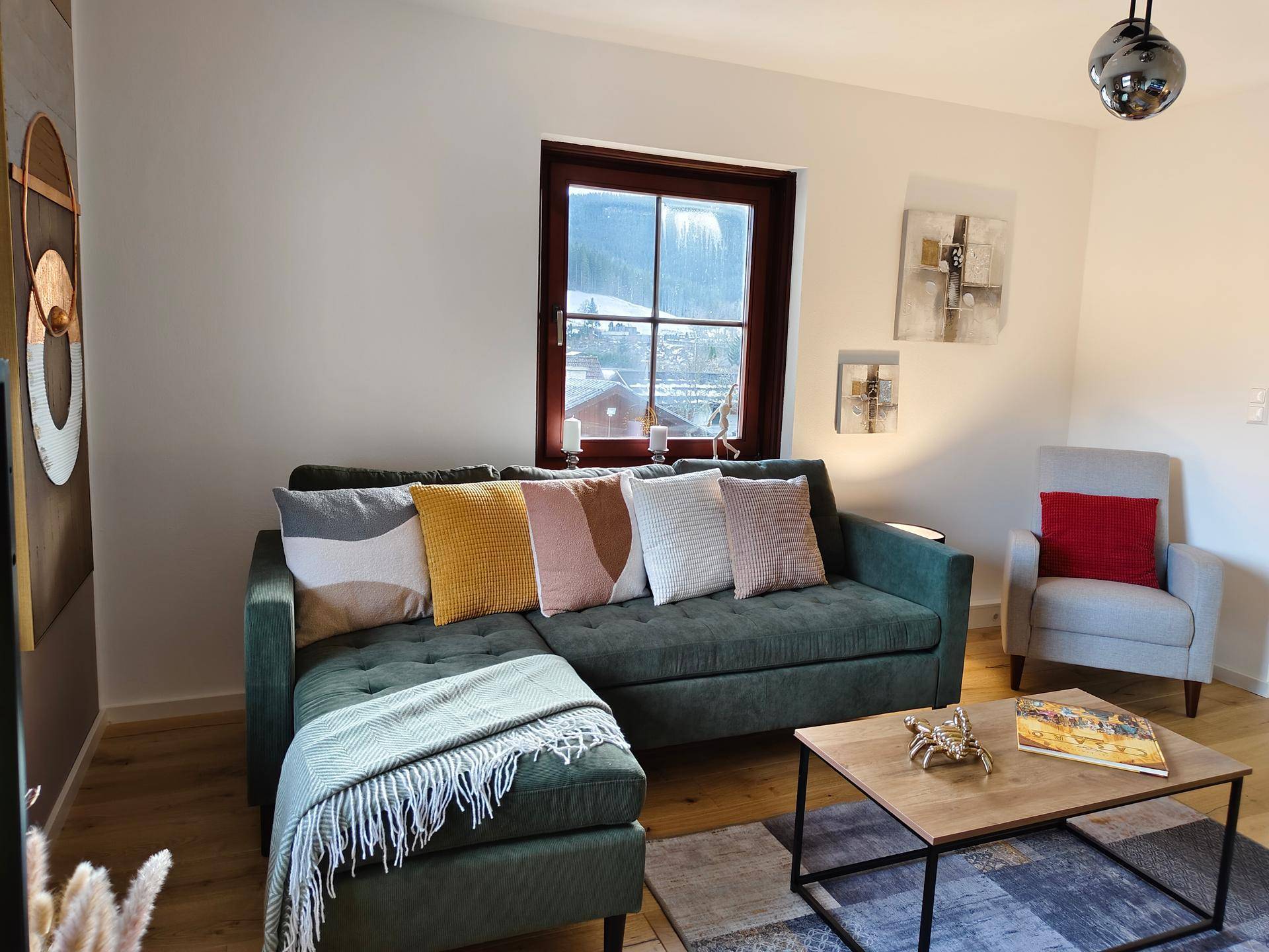 Gemütliches Wohnzimmer mit grüner Eckcouch, bunten Kissen und Blick auf die verschneite Landschaft.