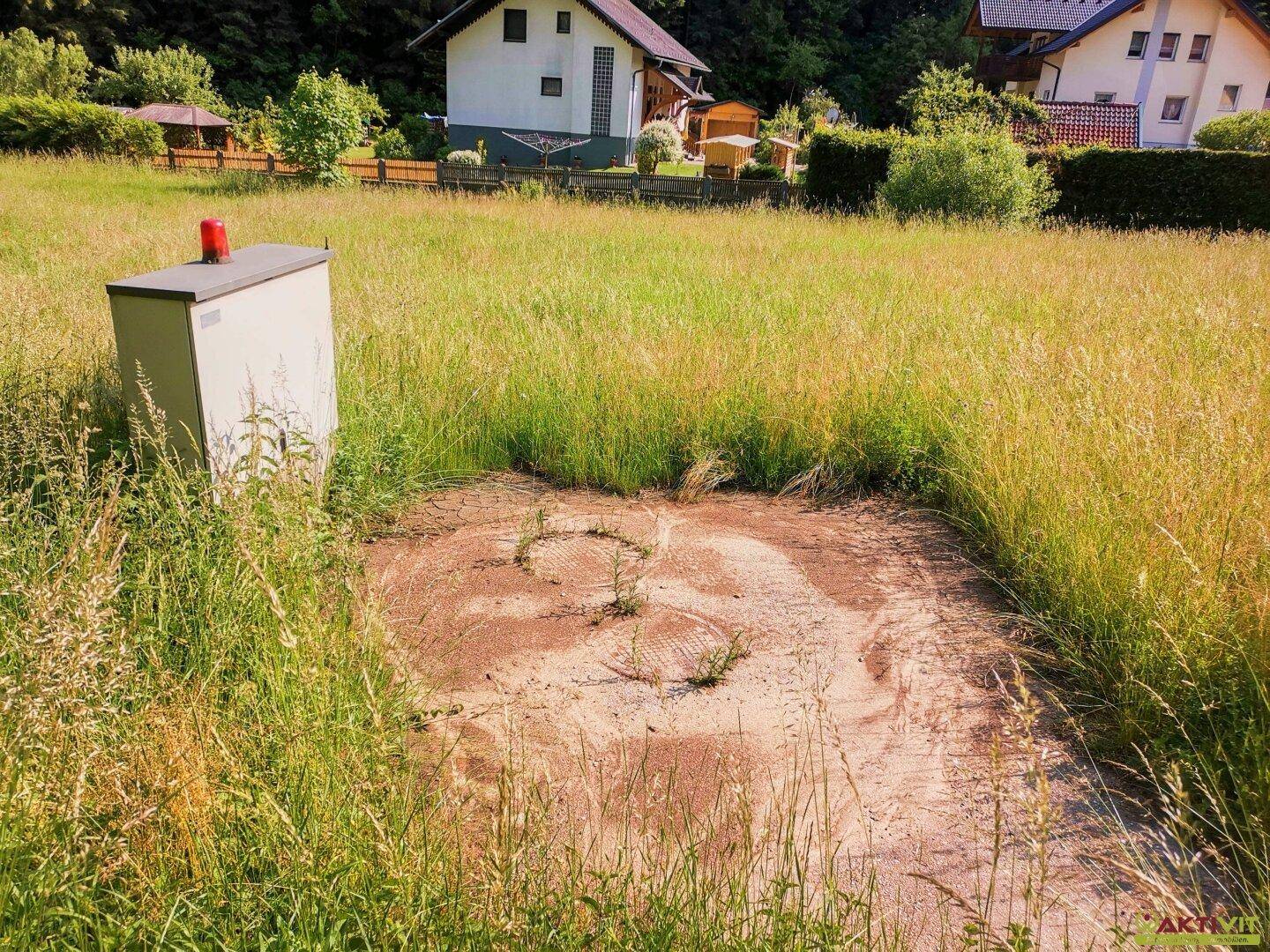 Grüne Wiese mit einem kleinen Versorgungskasten und Häusern im Hintergrund unter hellem Himmel.